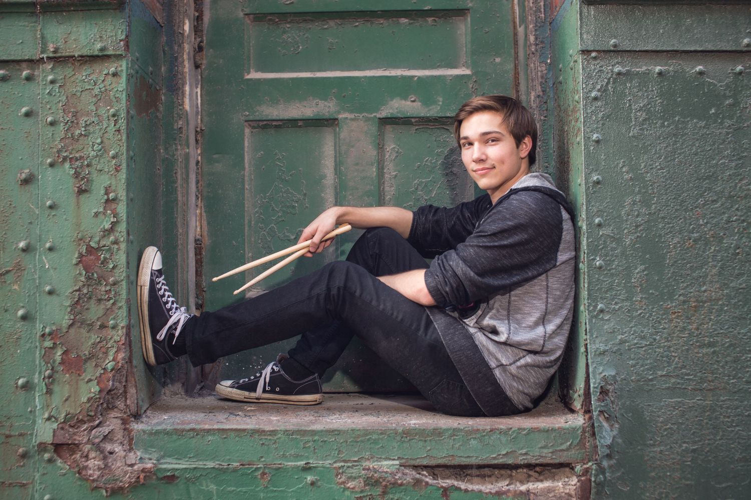 Young man sitting in a vintage green doorway holding drumsticks, wearing casual black outfit and sneakers.