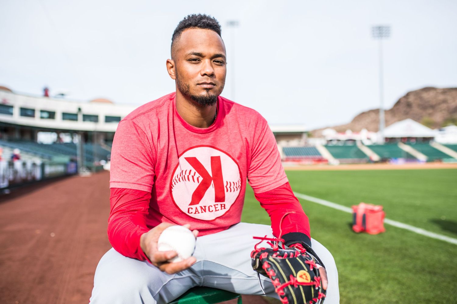 Baseball player in red jersey sits on field with glove and ball at stadium.