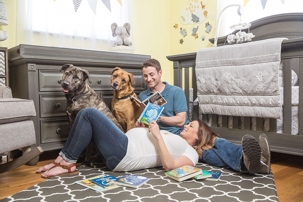 A couple relaxes on a gray patterned rug in a nursery room with their two dogs and baby.