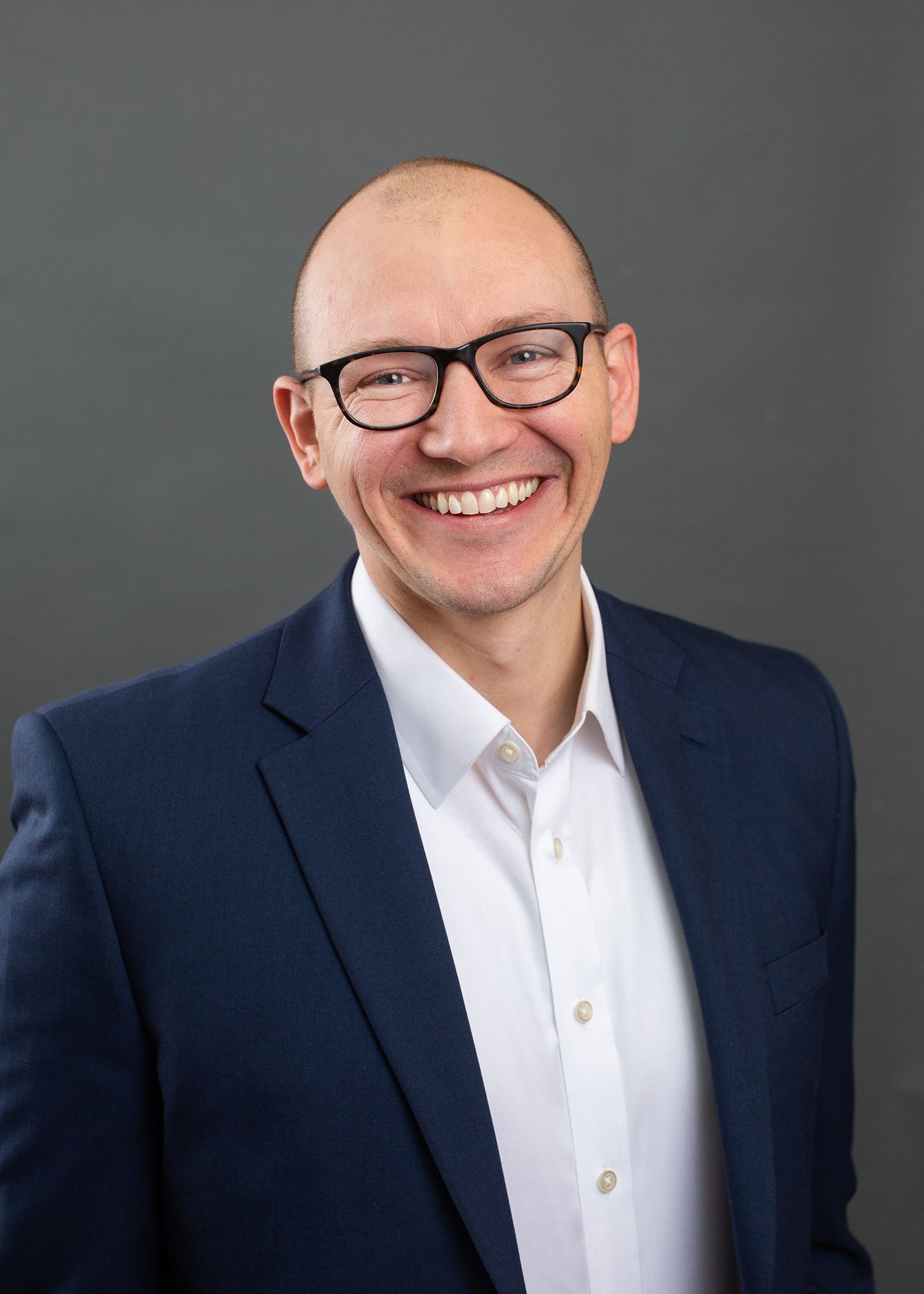 Professional in navy suit and glasses poses for headshot against grey background.