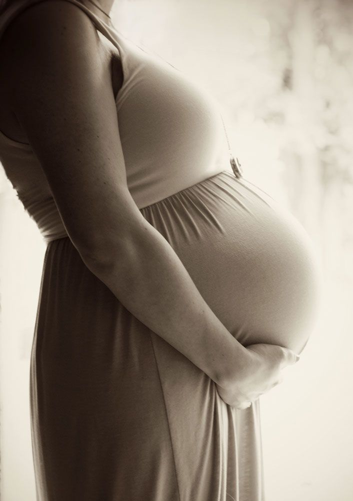 Black and white artistic portrait showing a pregnant woman in a long dress holding her baby bump tenderly.