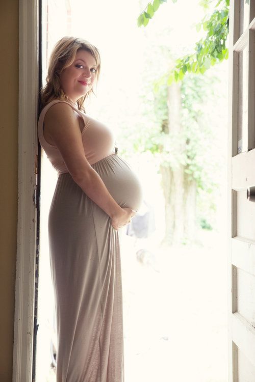 Maternity portrait of a pregnant woman in a long gray dress by a sunlit window.