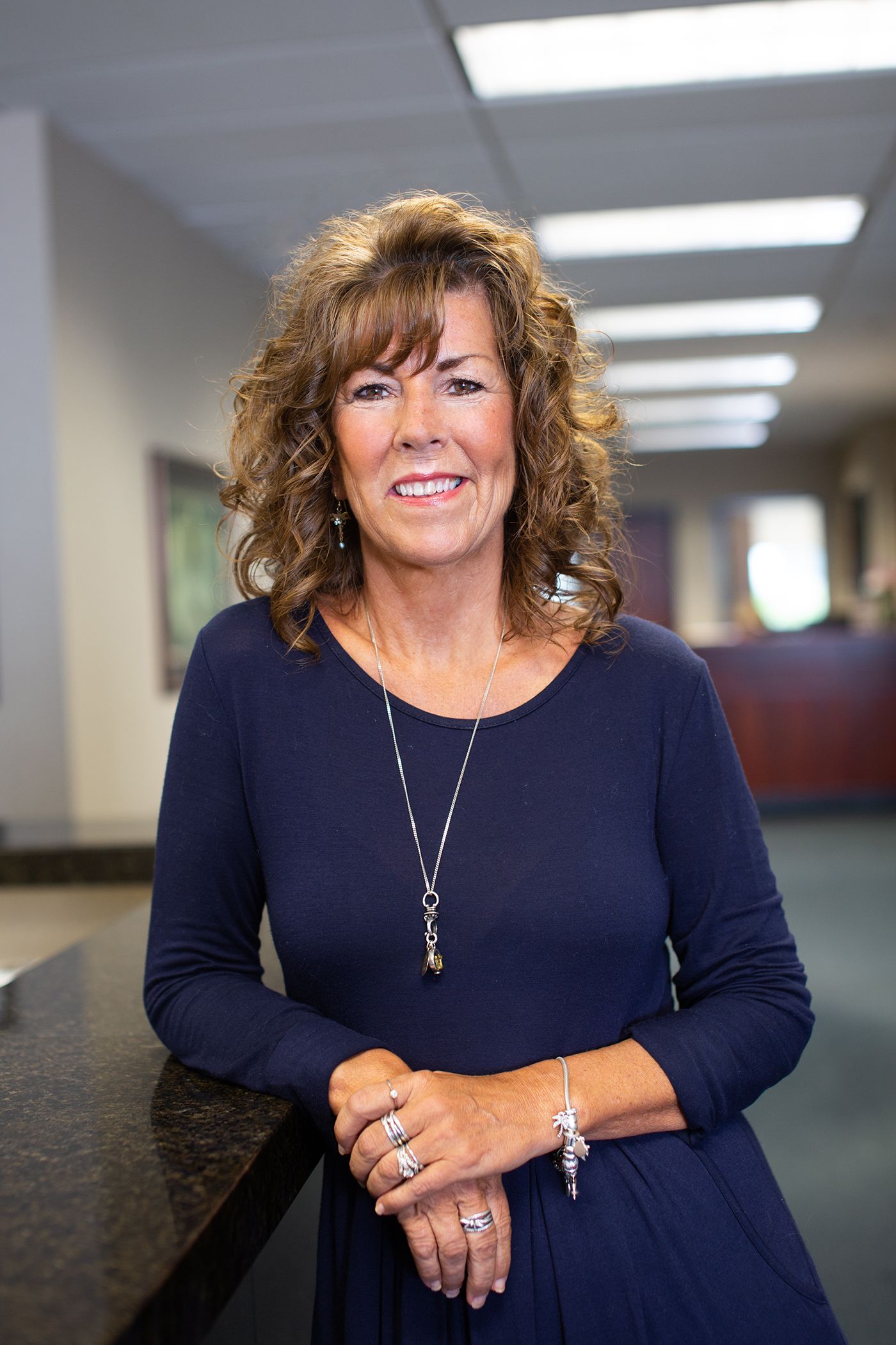 Professional woman wearing navy dress and necklace smiles warmly in office hallway setting.