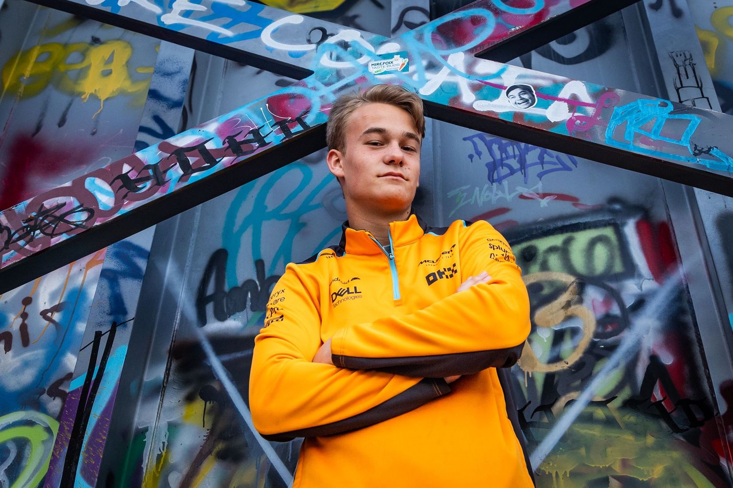 Young man in orange McLaren F1 team jacket poses confidently with arms crossed in front of colorful graffiti wall.