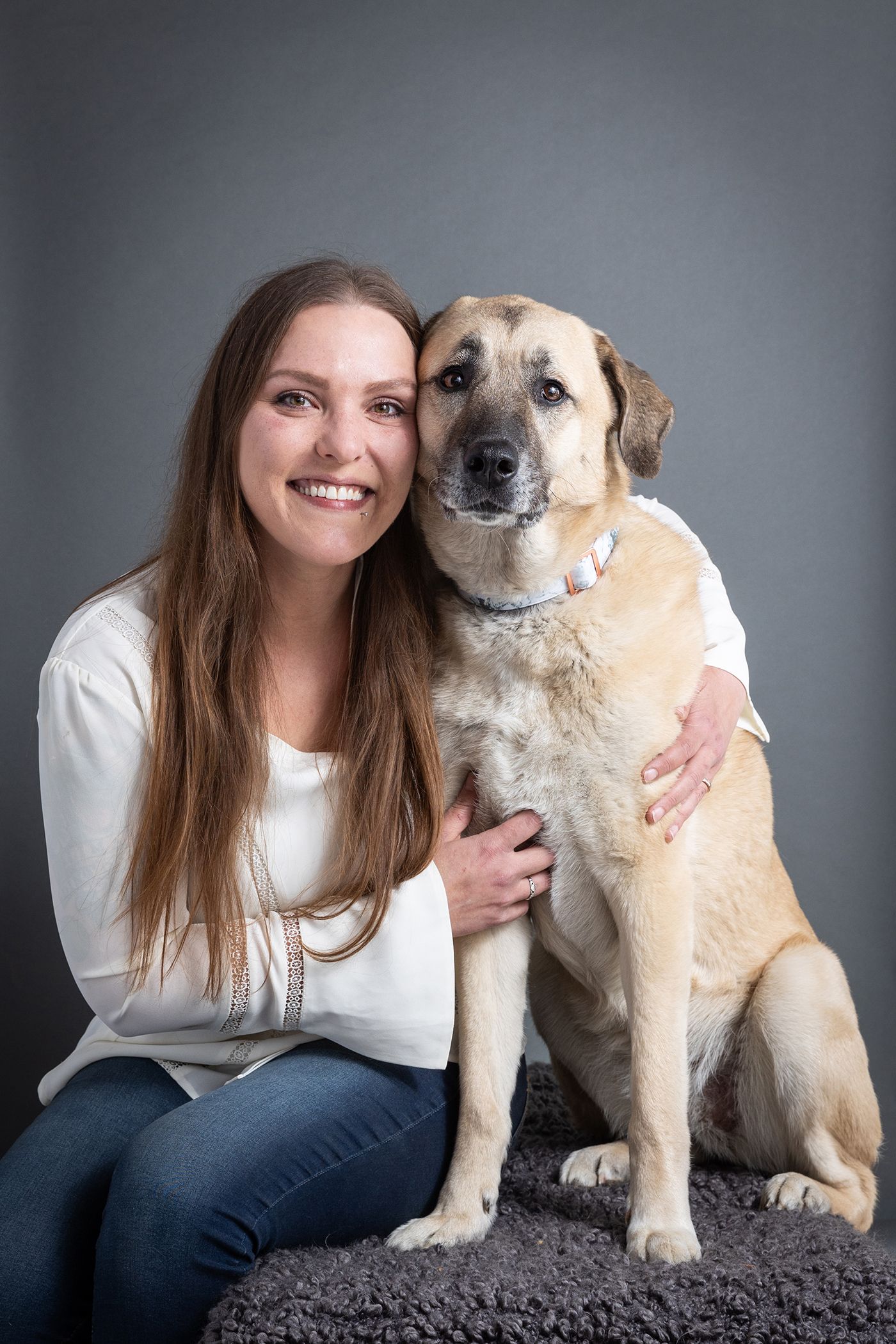 Smiling woman with long brown hair in white top hugs a large tan dog while sitting on gray carpet.