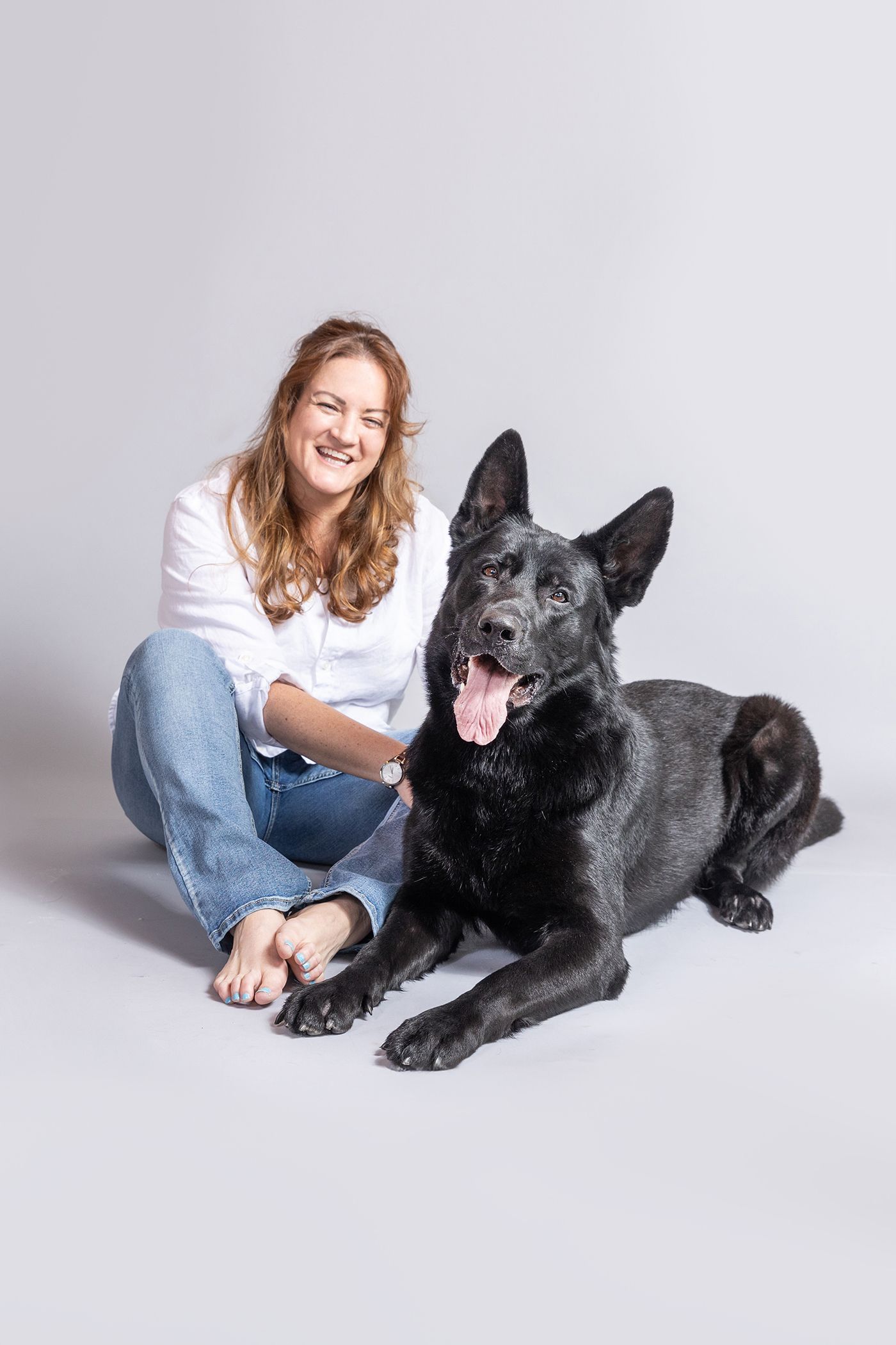 Smiling woman in white shirt and jeans sitting on floor with large black German Shepherd dog in studio.