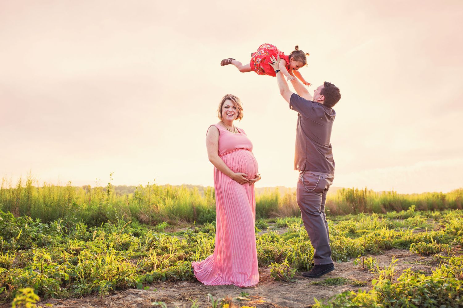 Family joyfully poses in a sunset meadow with pink maternity gown as father lifts child playfully in the air.