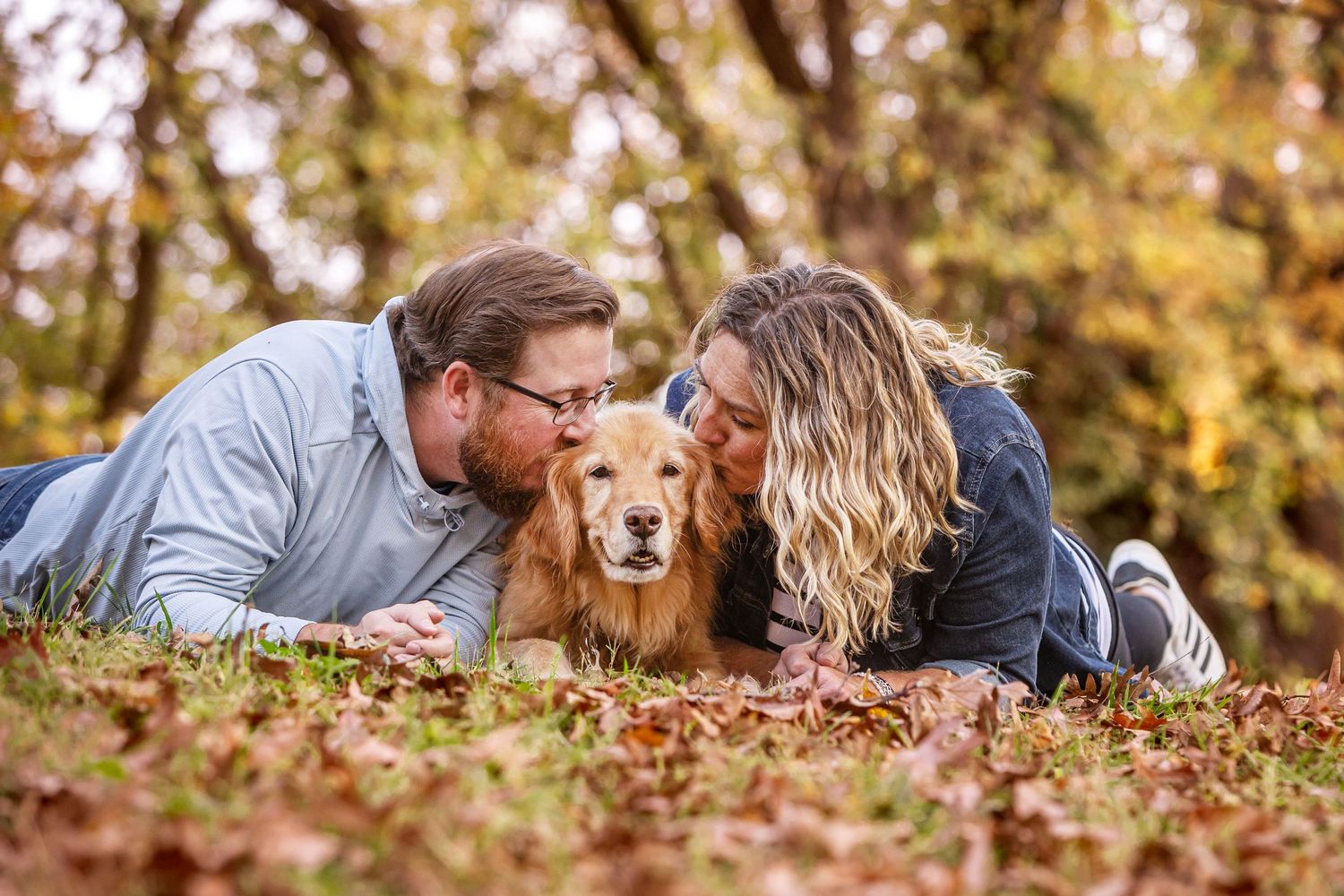 Couple lying on autumn leaves kissing their golden retriever dog in a fall forest setting.