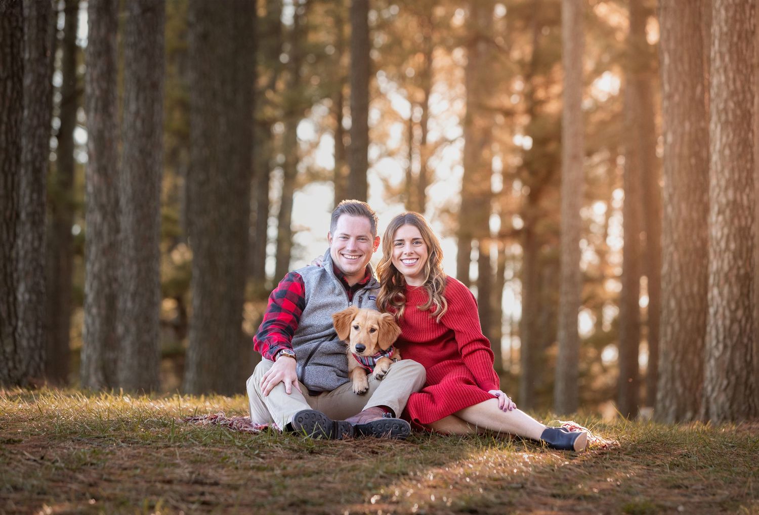 A couple sits with their small dog in a sunlit forest during autumn, wearing red and white casual attire.