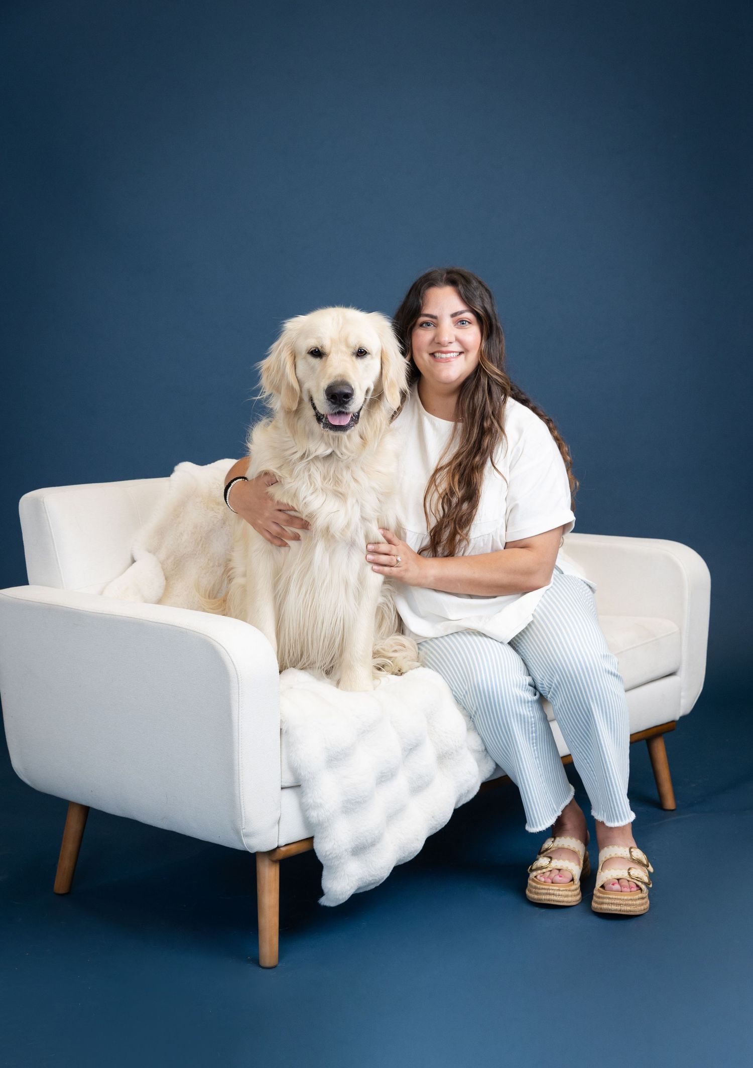 Woman smiling with golden retriever on white sofa against blue background in professional pet portrait.