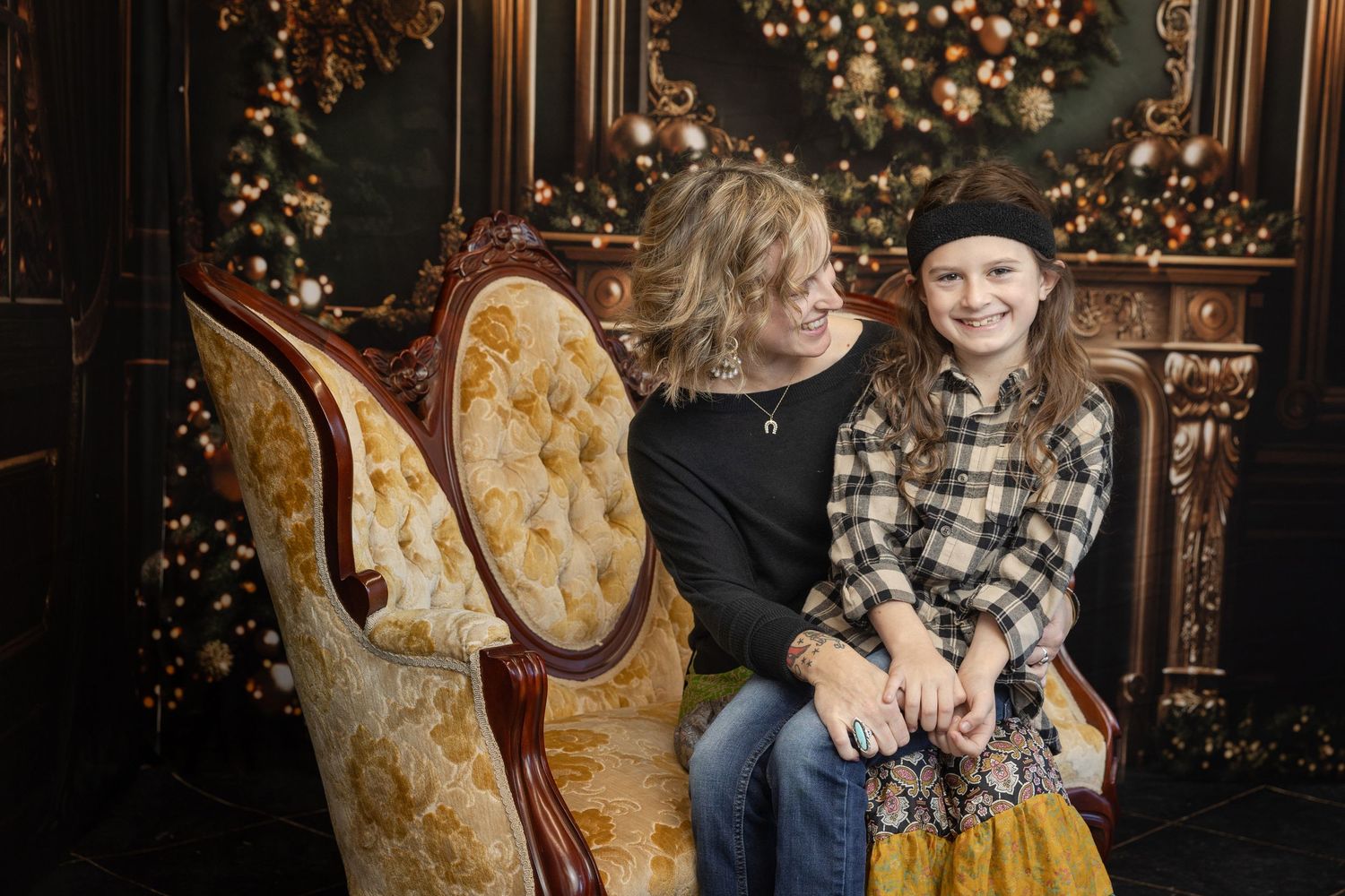 Woman and young girl smiling together on ornate gold chair with festive Christmas decorations backdrop.