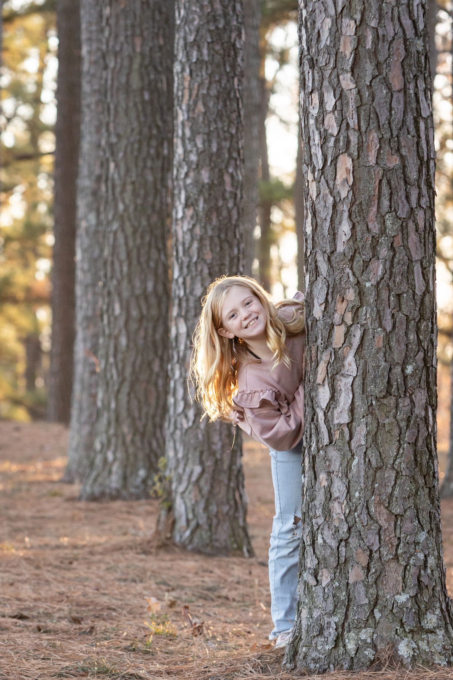 Young girl with long blonde hair peeking around a pine tree trunk in a forest, smiling, wearing pink top and jeans.
