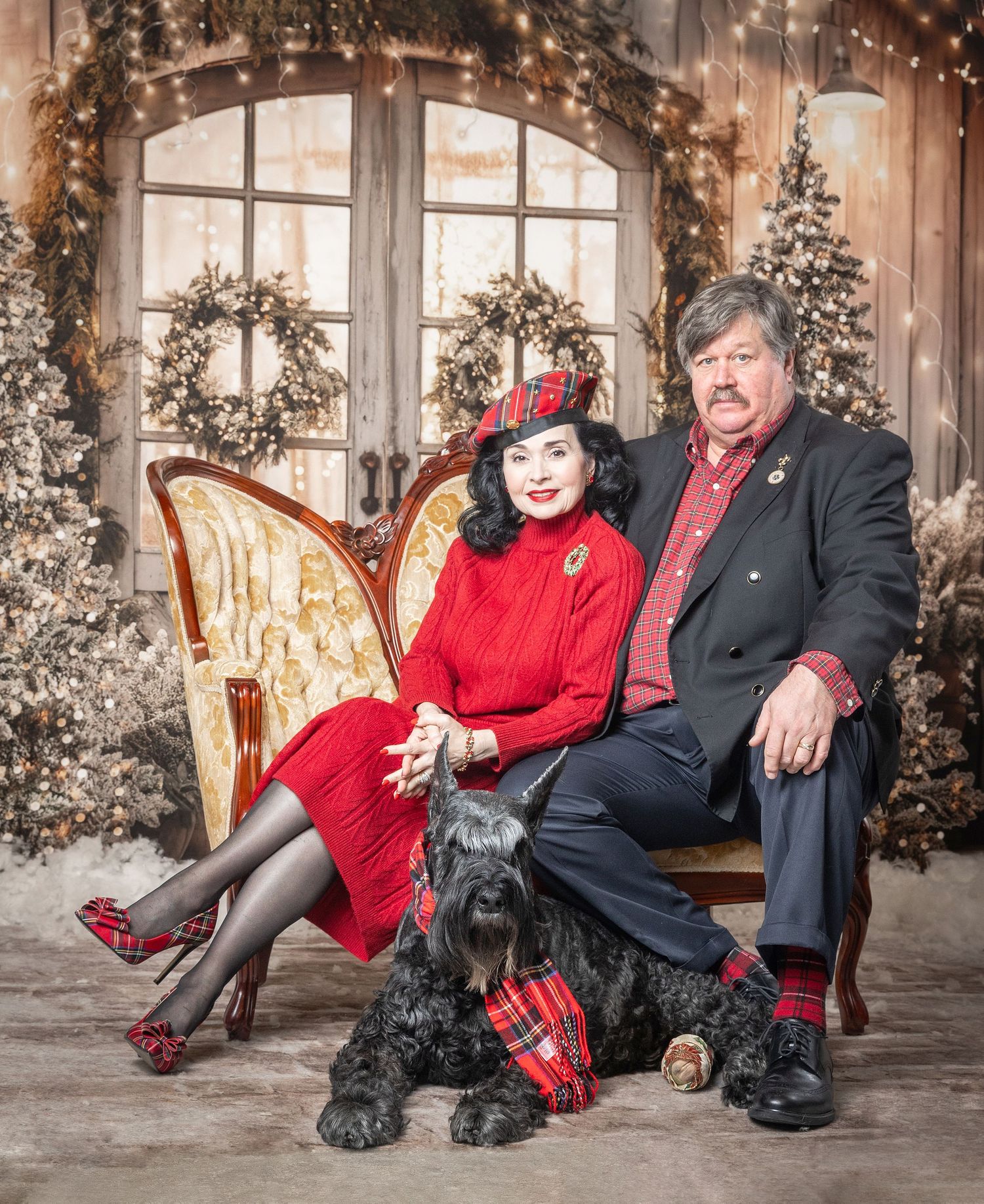 Elegant couple in matching red outfits pose with black Scottish Terrier before festive Christmas backdrop.