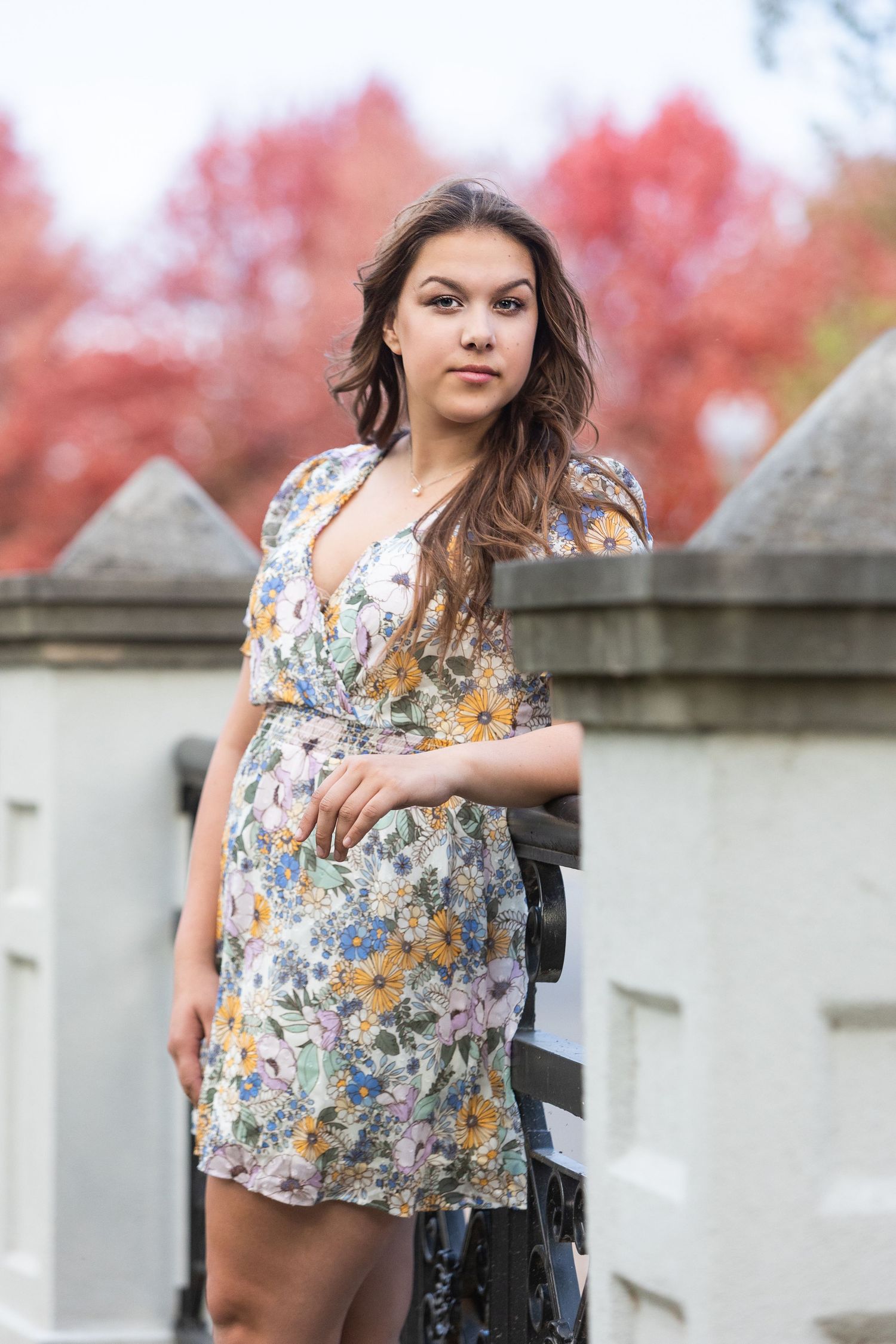 Young woman in floral dress poses by stone fence with vibrant red autumn foliage in background.