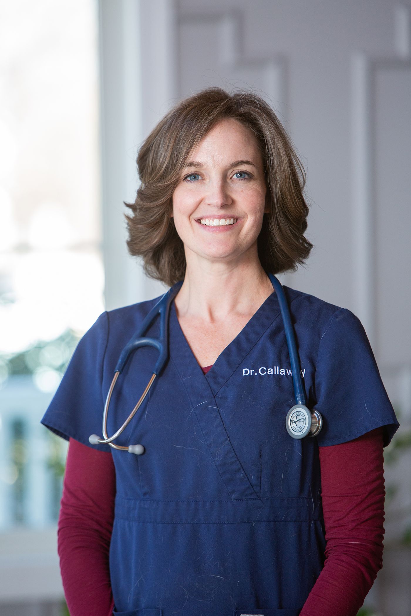 Healthcare professional wearing navy scrubs and stethoscope poses in medical office setting.