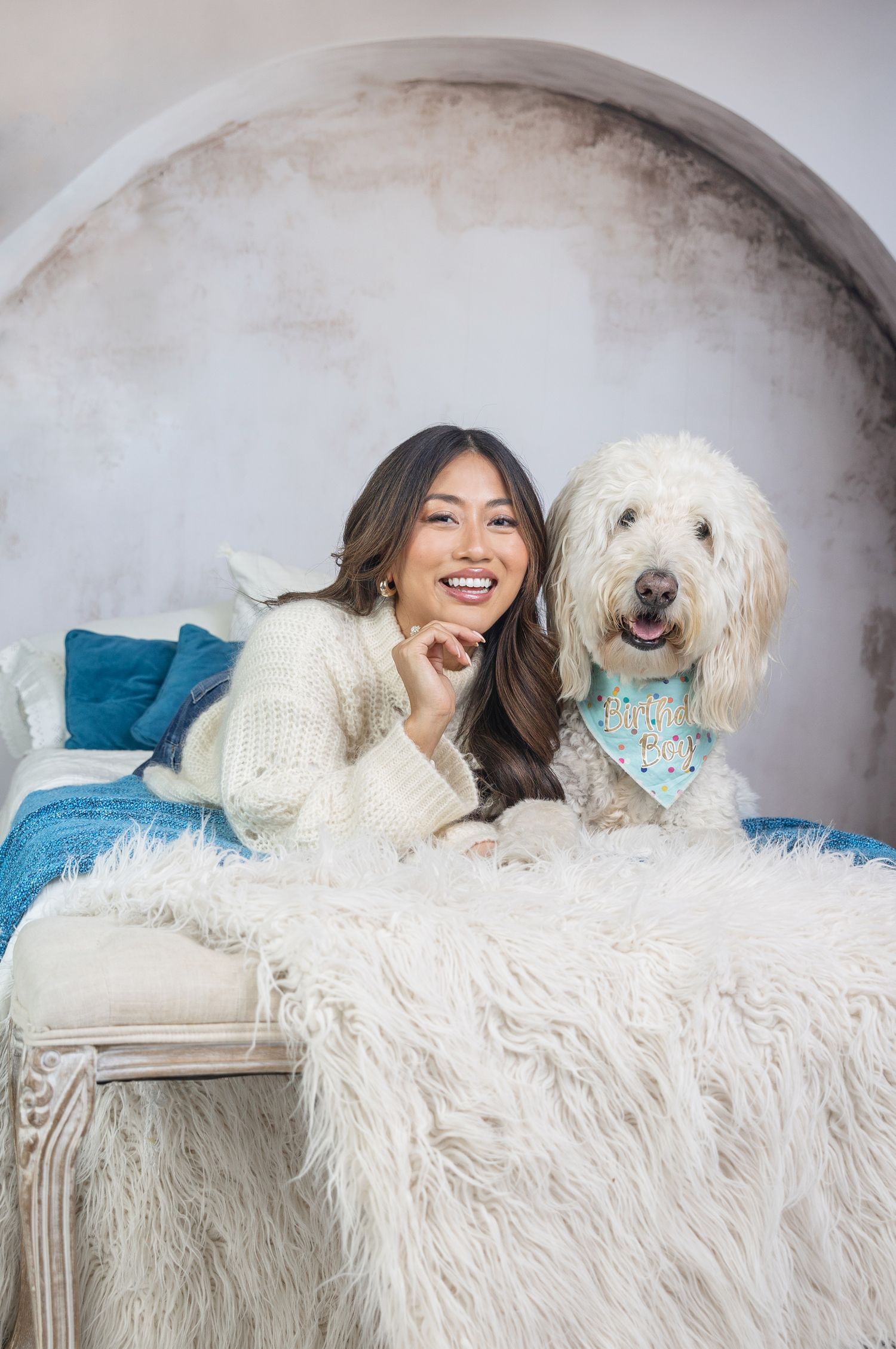 Smiling woman in cream sweater posing with fluffy white Goldendoodle wearing blue bandana on fur blanket.