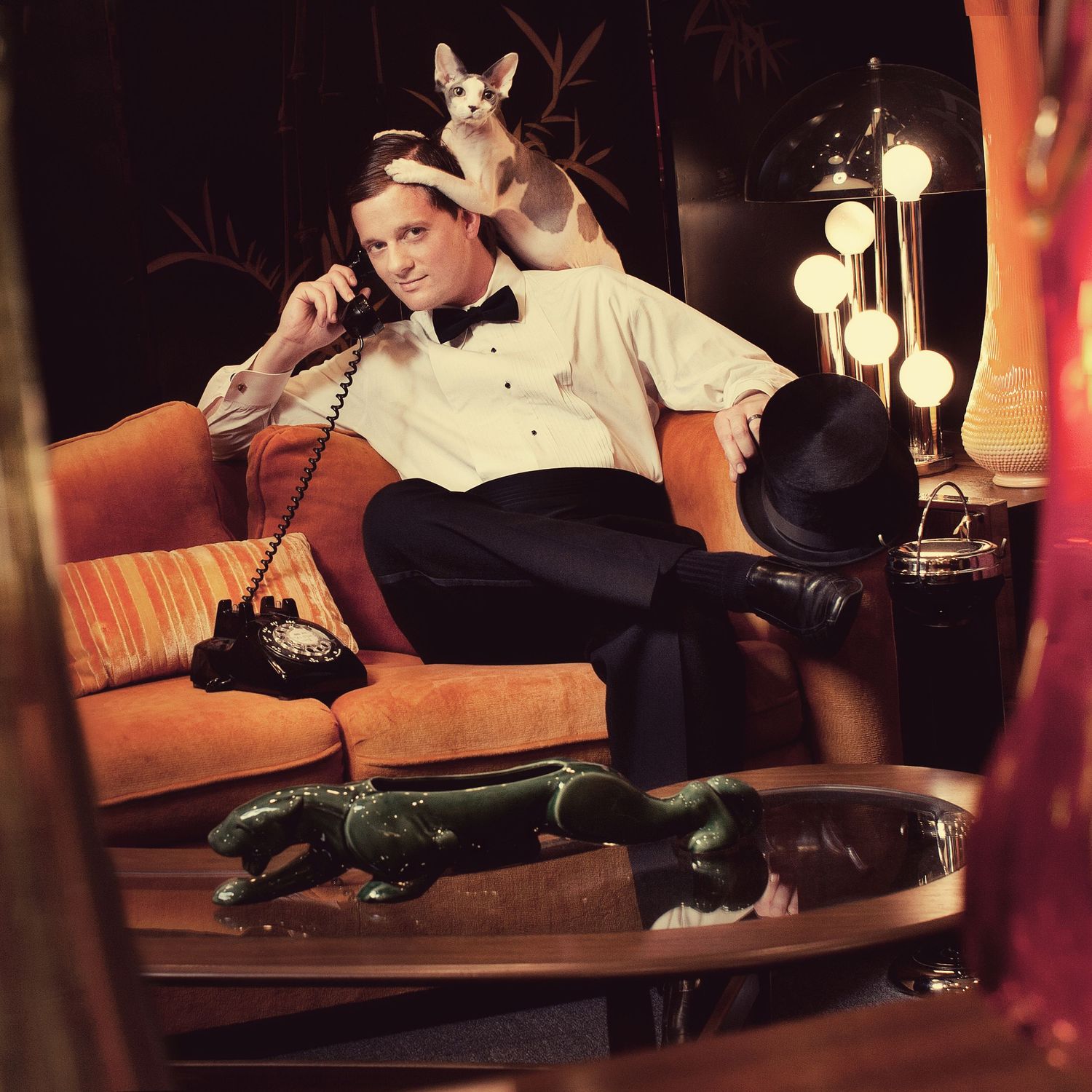 Retro-styled scene with someone in formal attire on orange couch with small dog and vintage phone.