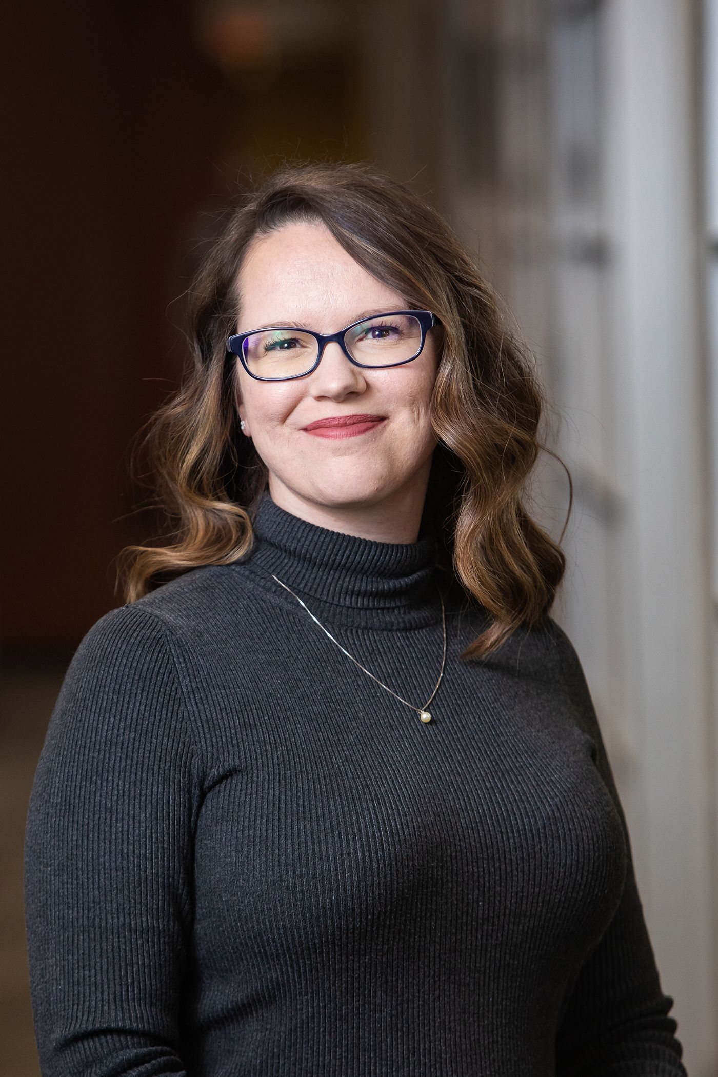 Professional headshot of person wearing black turtleneck sweater and glasses in office setting.