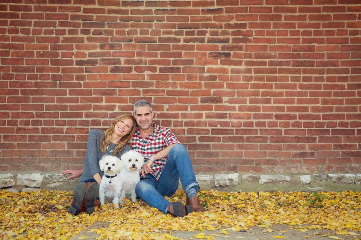 A couple sits with their two small white dogs against a brick wall surrounded by yellow autumn leaves.