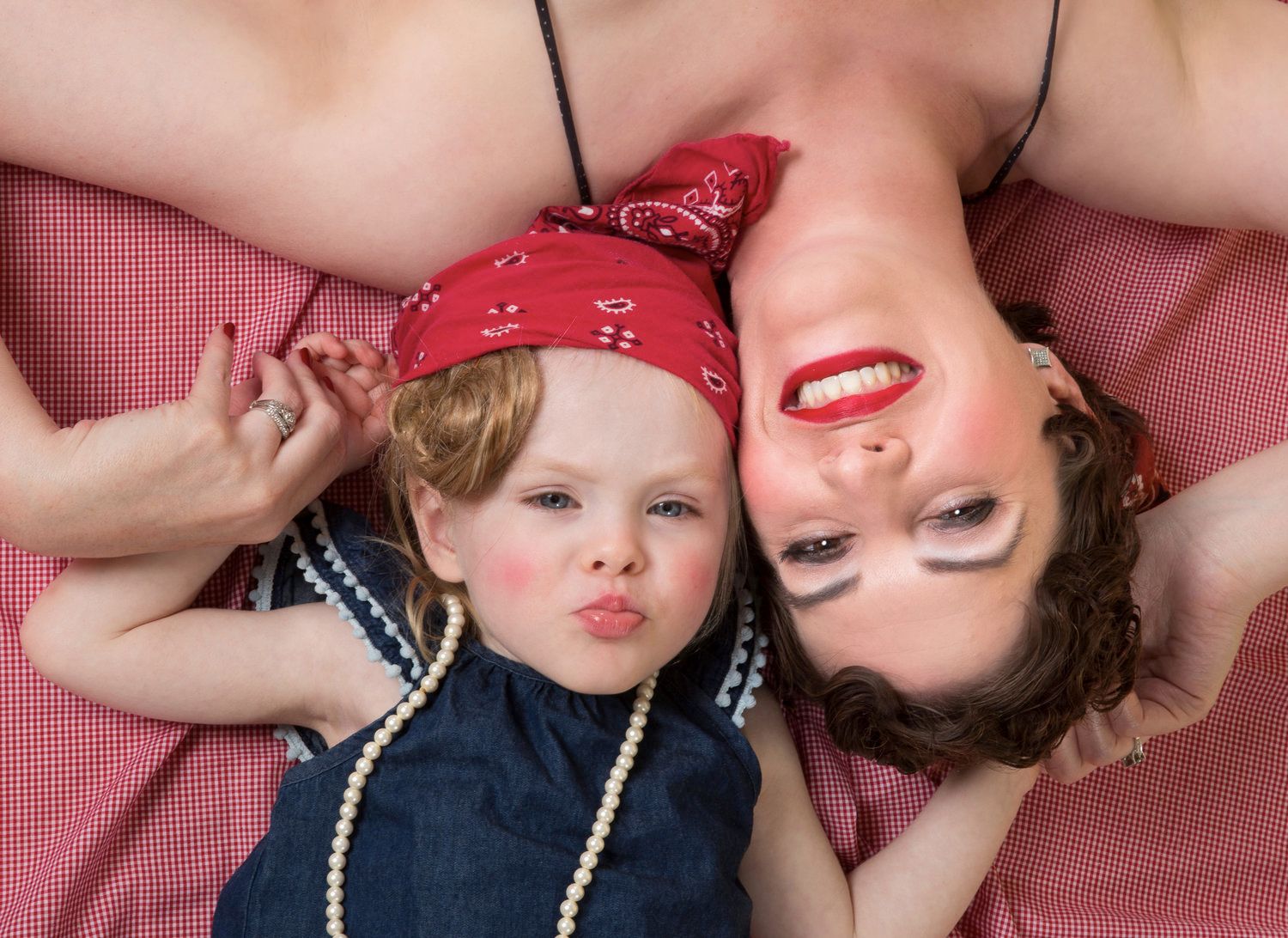 Mother and child lying on pink bedding wearing pearls and bandanas while making silly faces.