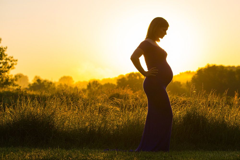 A silhouette of an expectant mother stands against a golden sunset in a field.