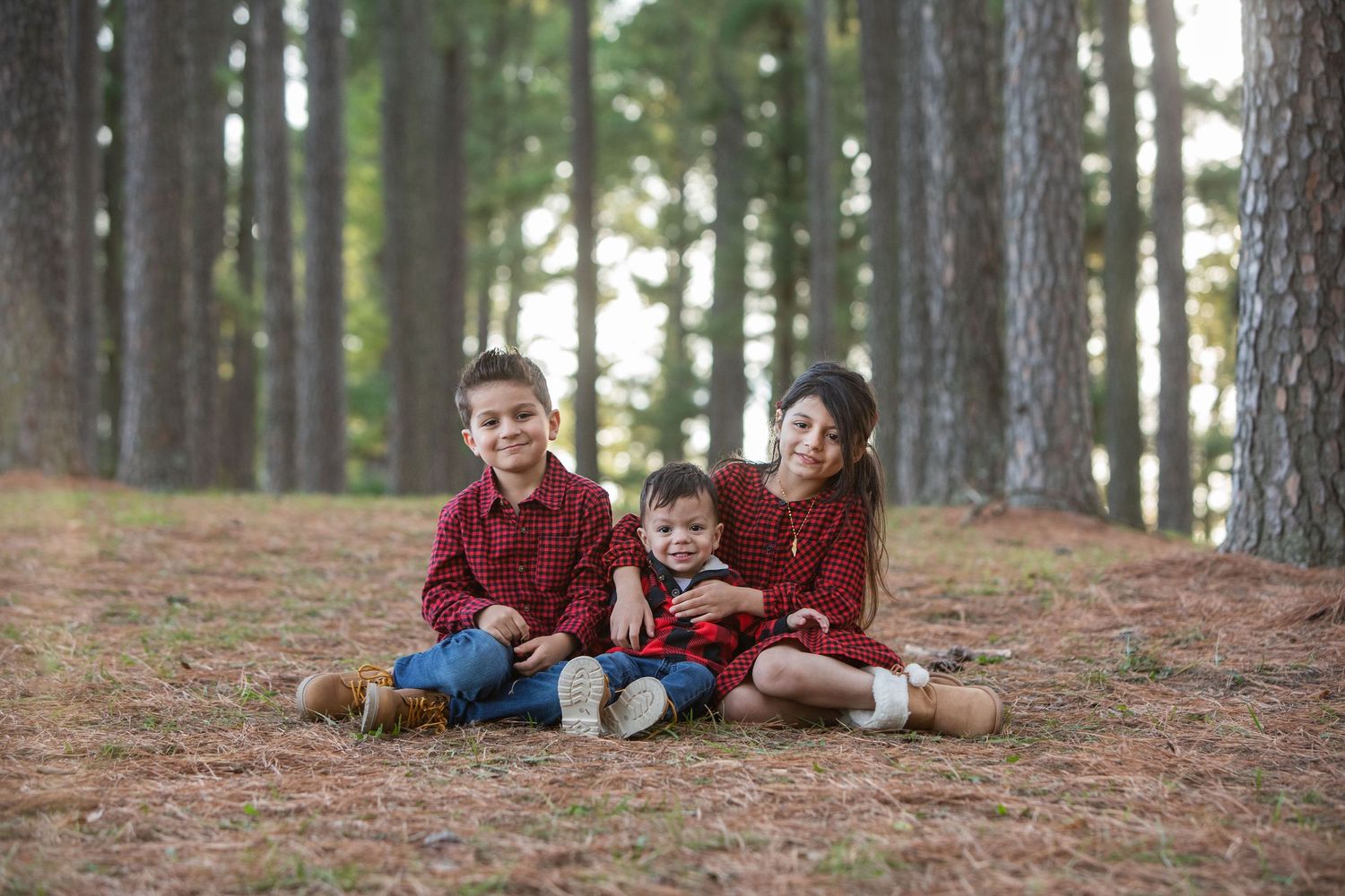 Three children in red outfits sit together among tall pine trees in a forest setting.