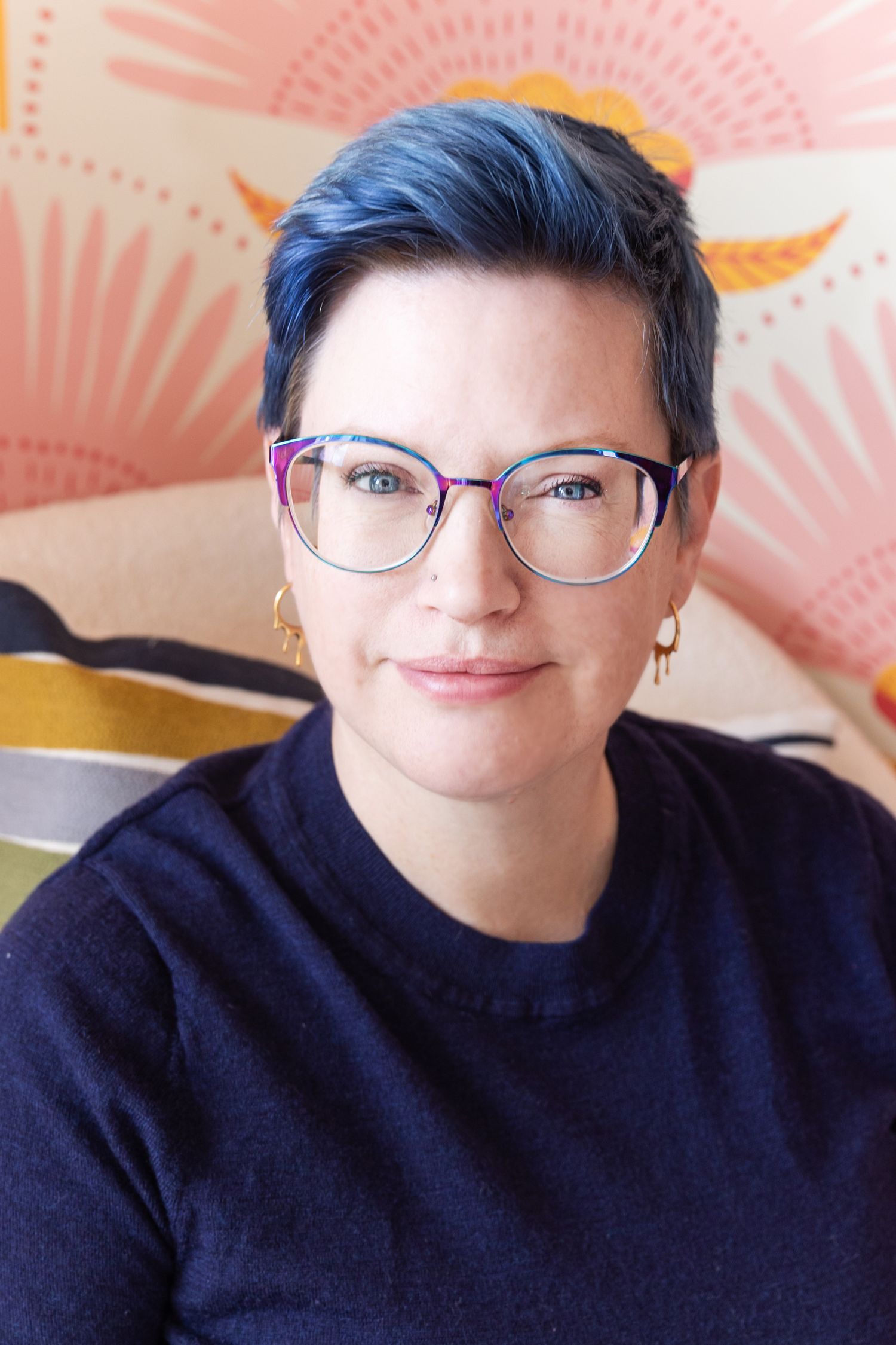 Person with blue hair wearing glasses and navy sweater smiles against colorful patterned background.