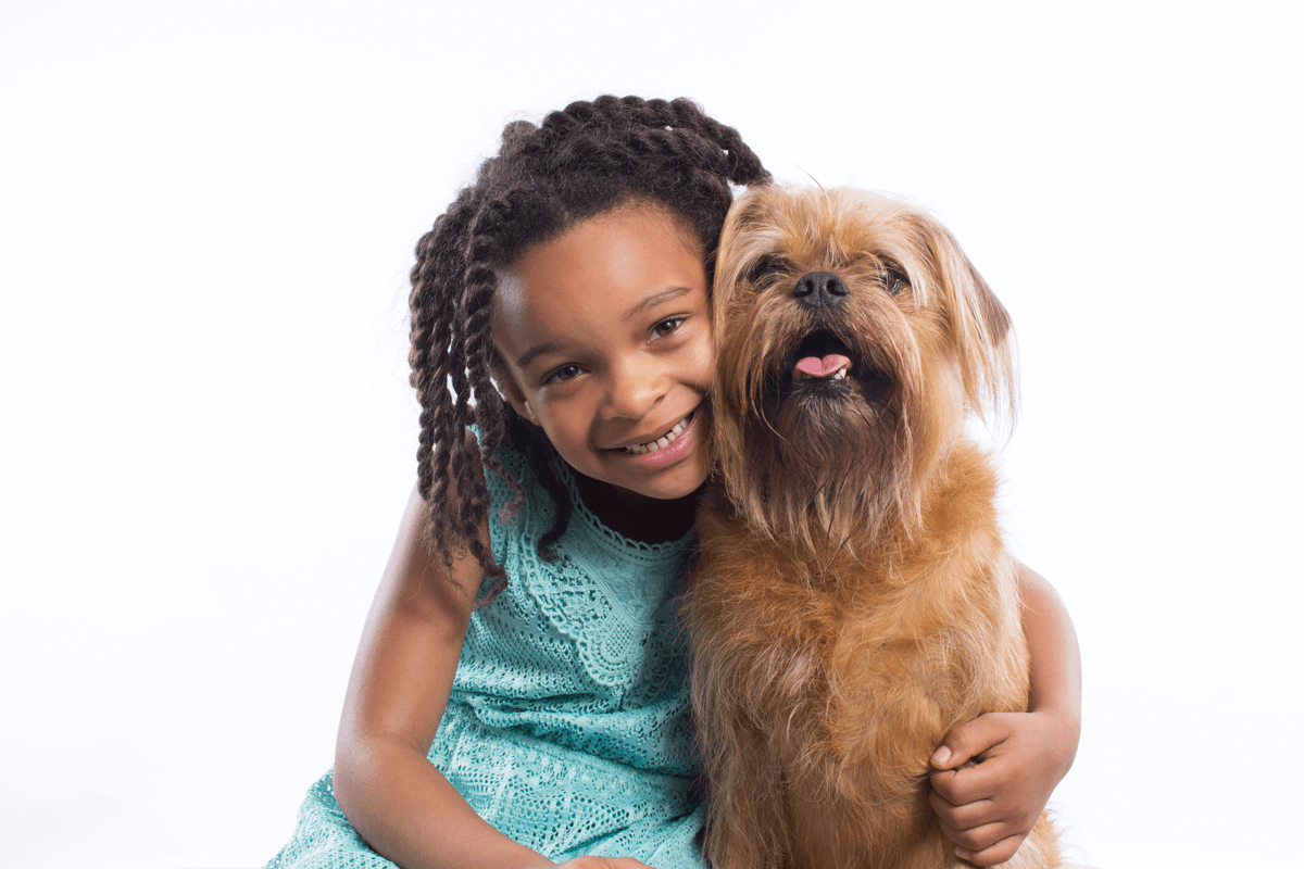 Young girl in turquoise dress poses with shaggy brown terrier dog against white background.