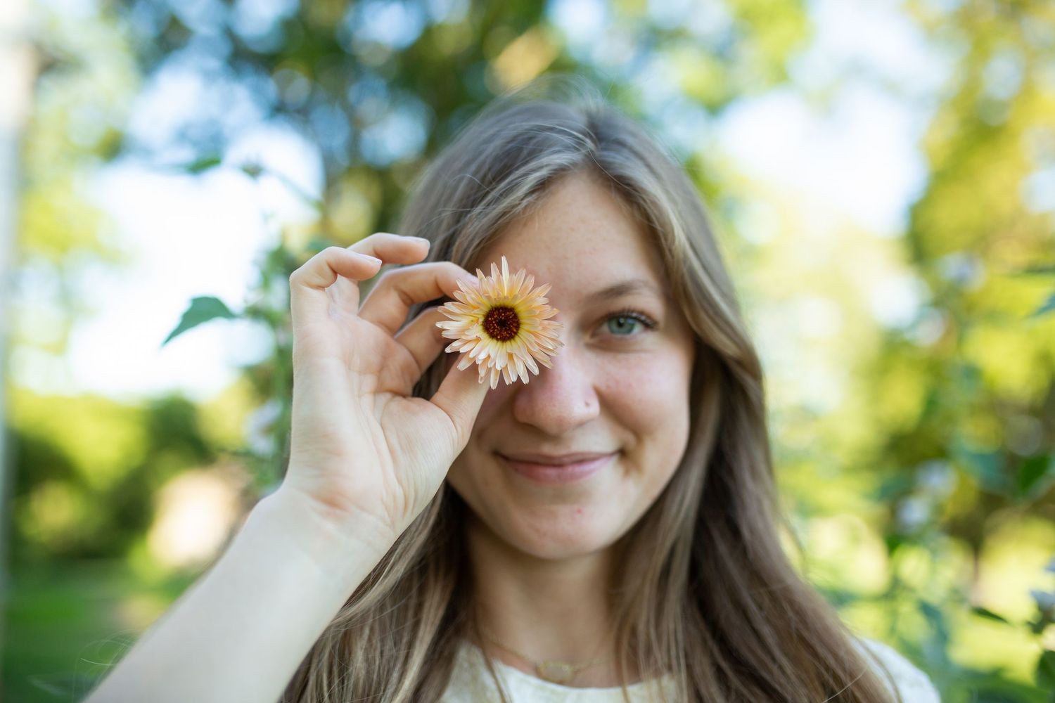 Young woman holding a yellow daisy flower over her eye, smiling outdoors in a green park setting.