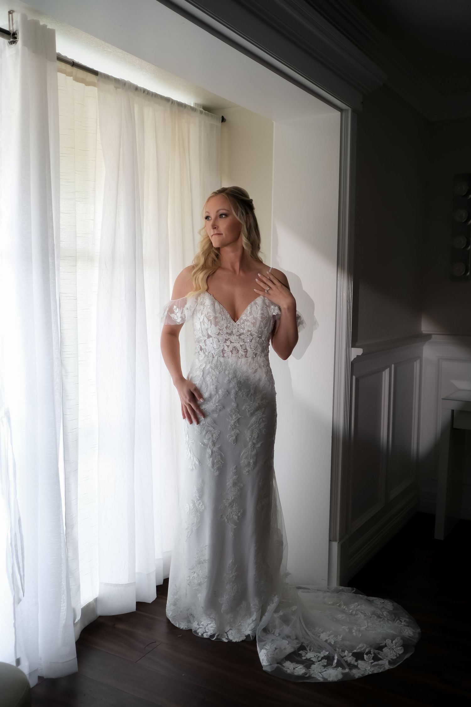 A bride stands by a window in an off-shoulder lace wedding gown with a flowing train.