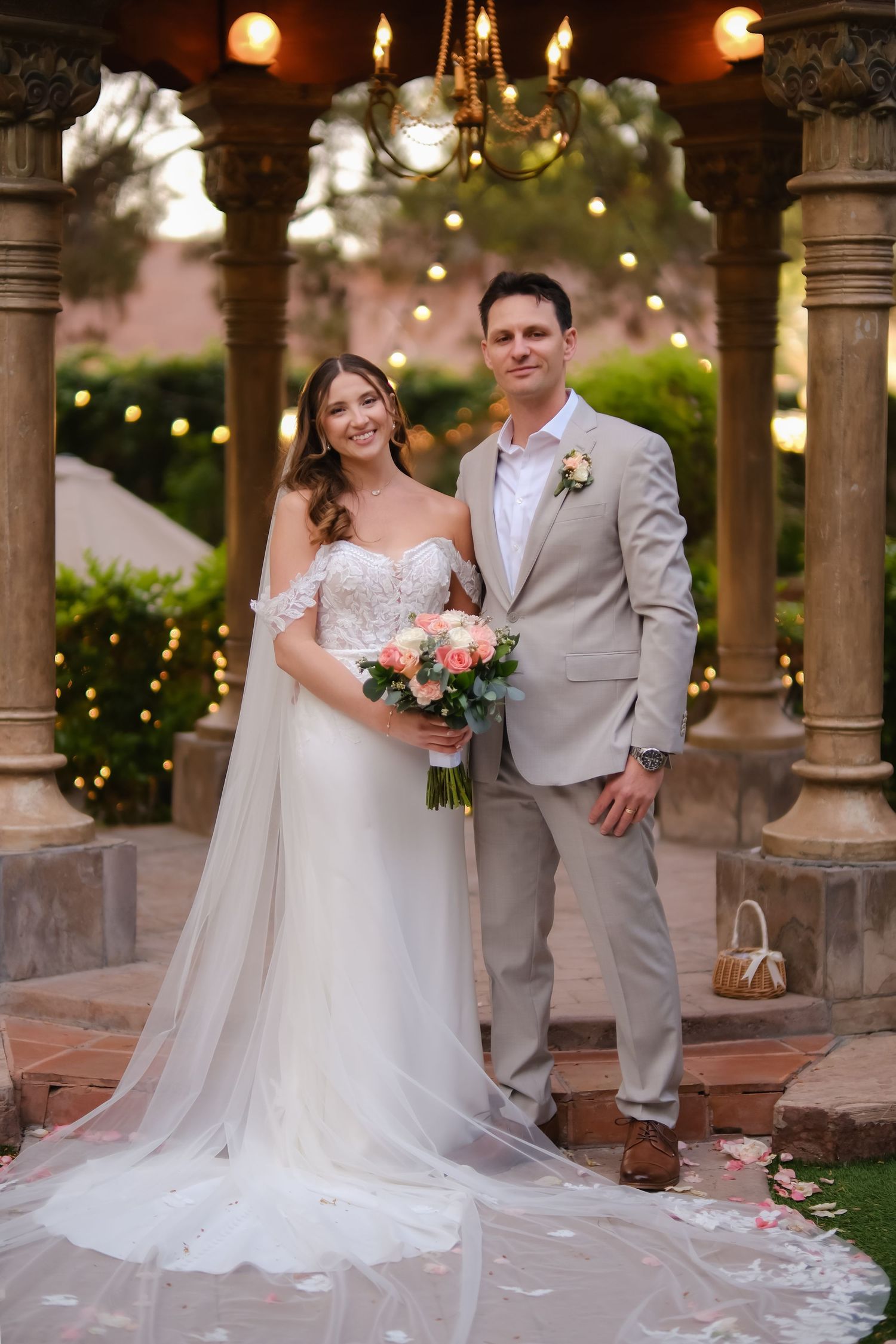 Elegant wedding couple standing under stone gazebo with string lights and floral decorations.