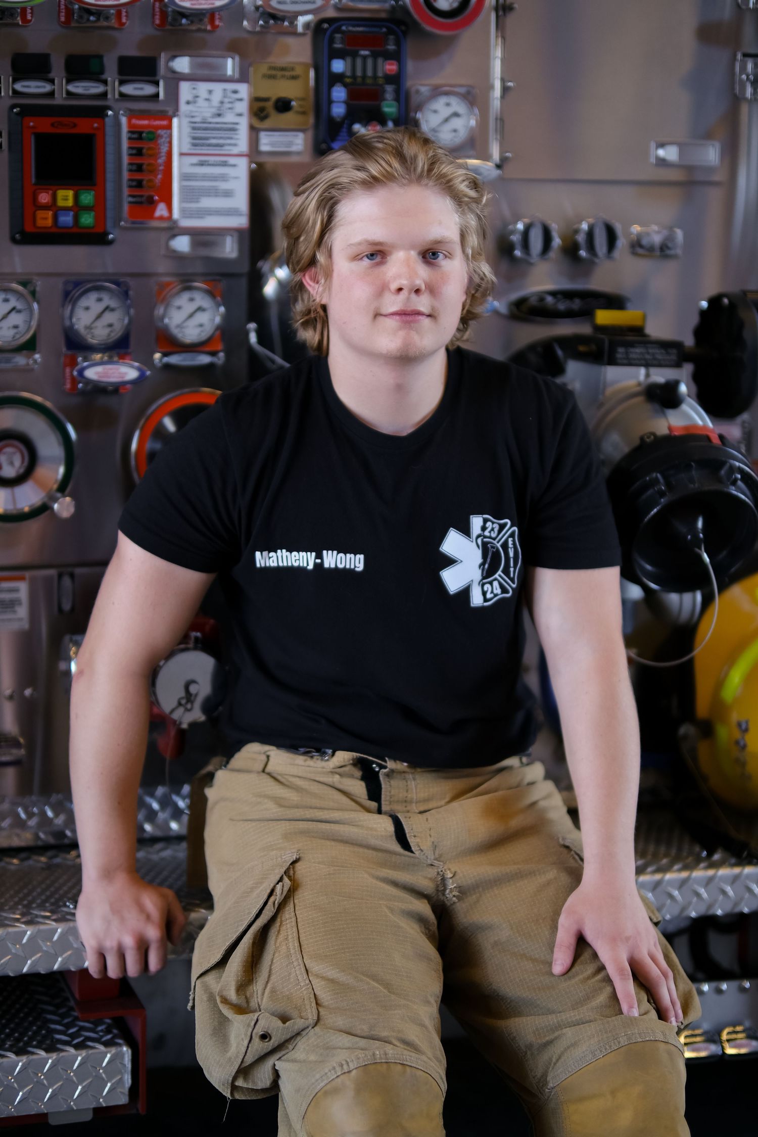EMT worker in black uniform shirt sits on equipment storage panel with various gauges and controls visible.