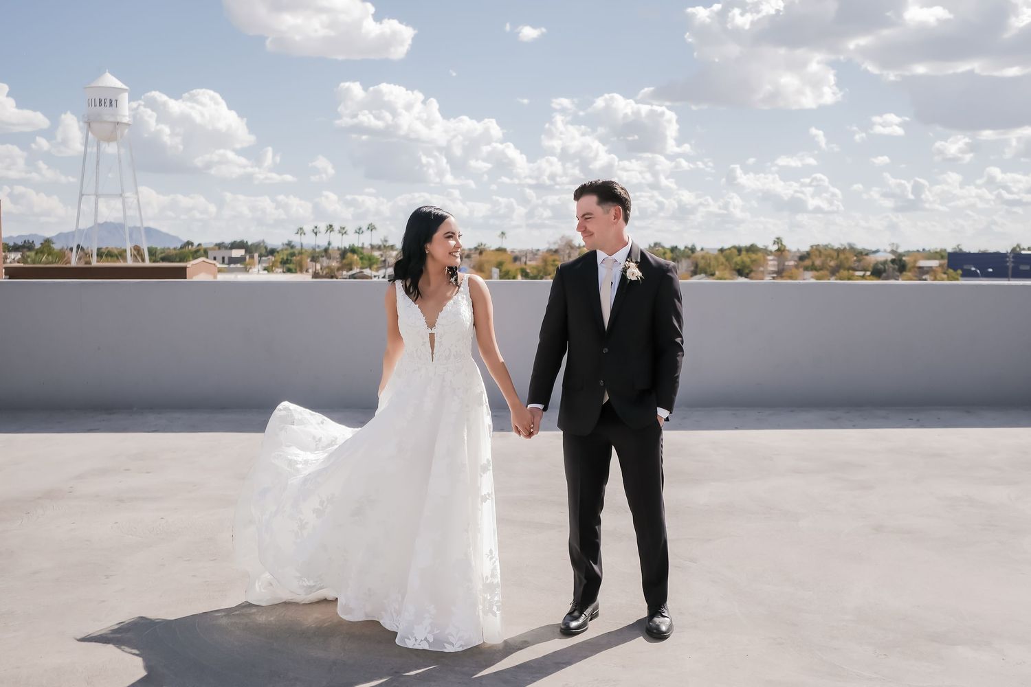 Bride and groom hold hands on rooftop under cloudy blue sky, bride in flowing white gown, groom in black tuxedo.