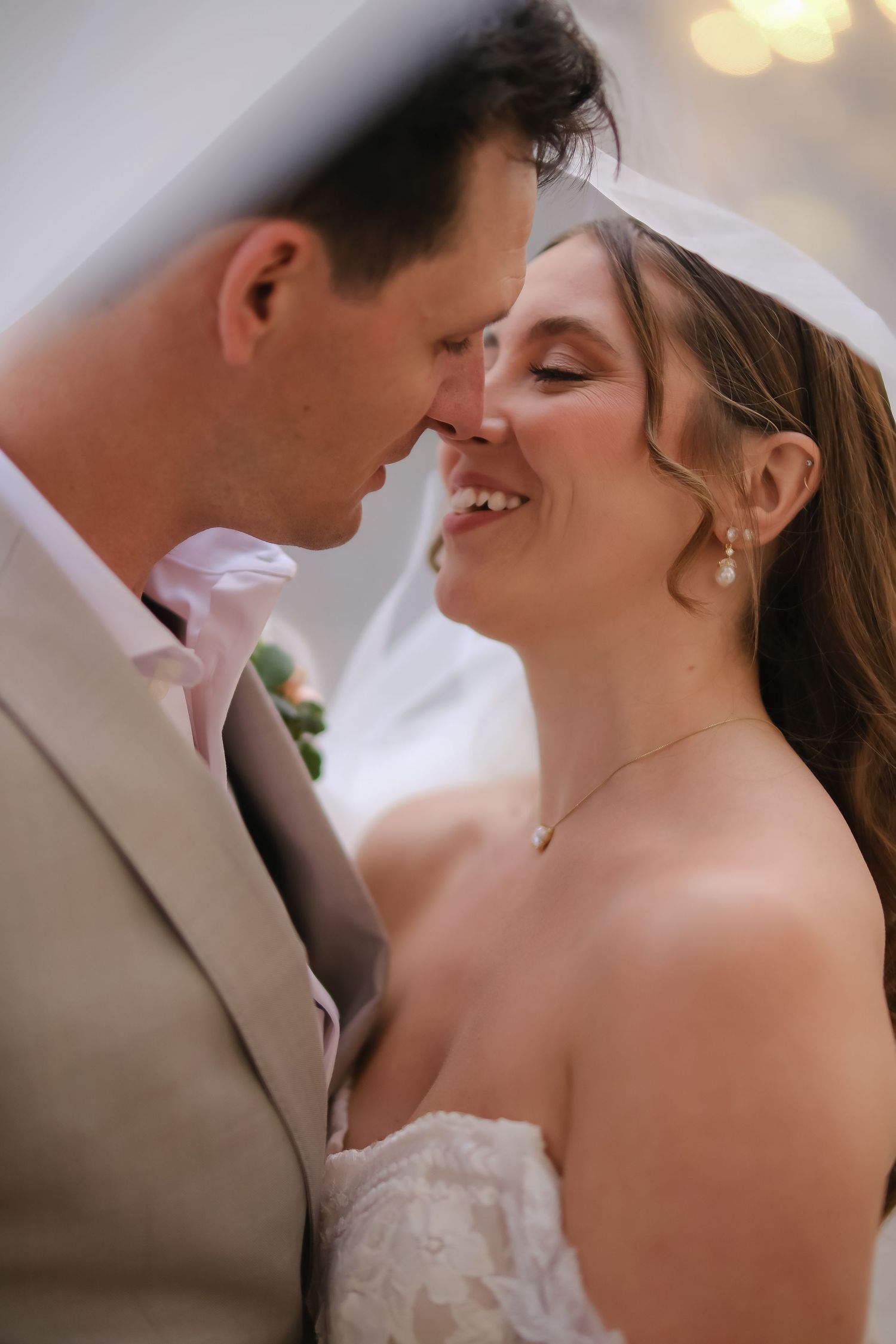 A joyful bride in lace gown and veil shares an intimate moment with her groom in a gray suit at their wedding.