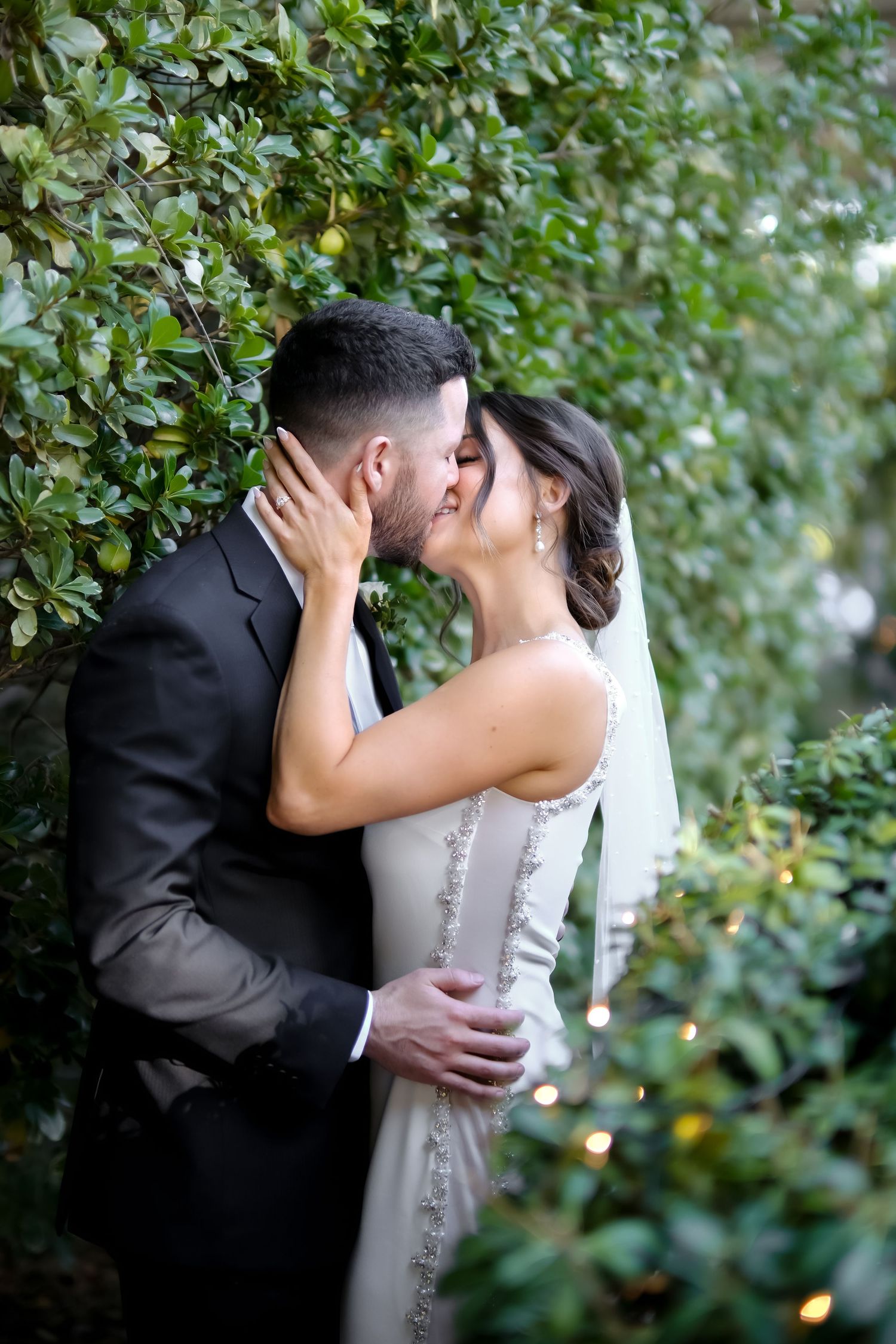 Newlyweds share intimate moment among garden greenery with soft natural lighting.