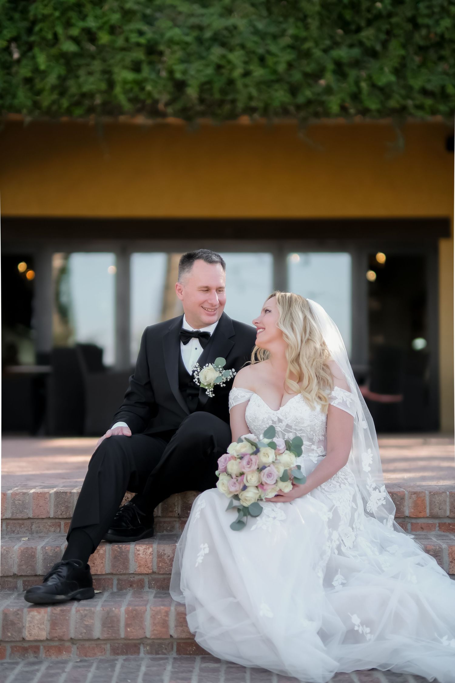 Bride and groom sitting on brick steps, smiling at each other, bride holding a pastel bouquet, groom in black tuxedo.