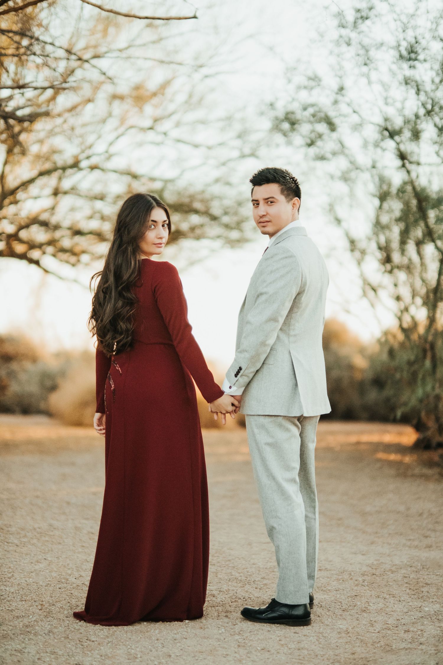 A couple in formal attire poses against a natural backdrop during golden hour with a burgundy dress and light suit.