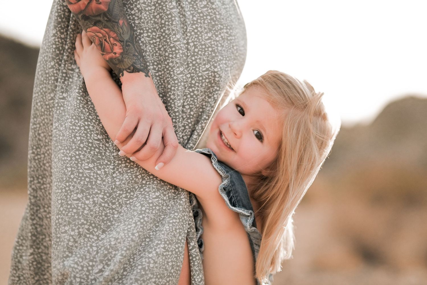 Playful toddler peeks out from behind someone wearing a grey dress in outdoor setting.