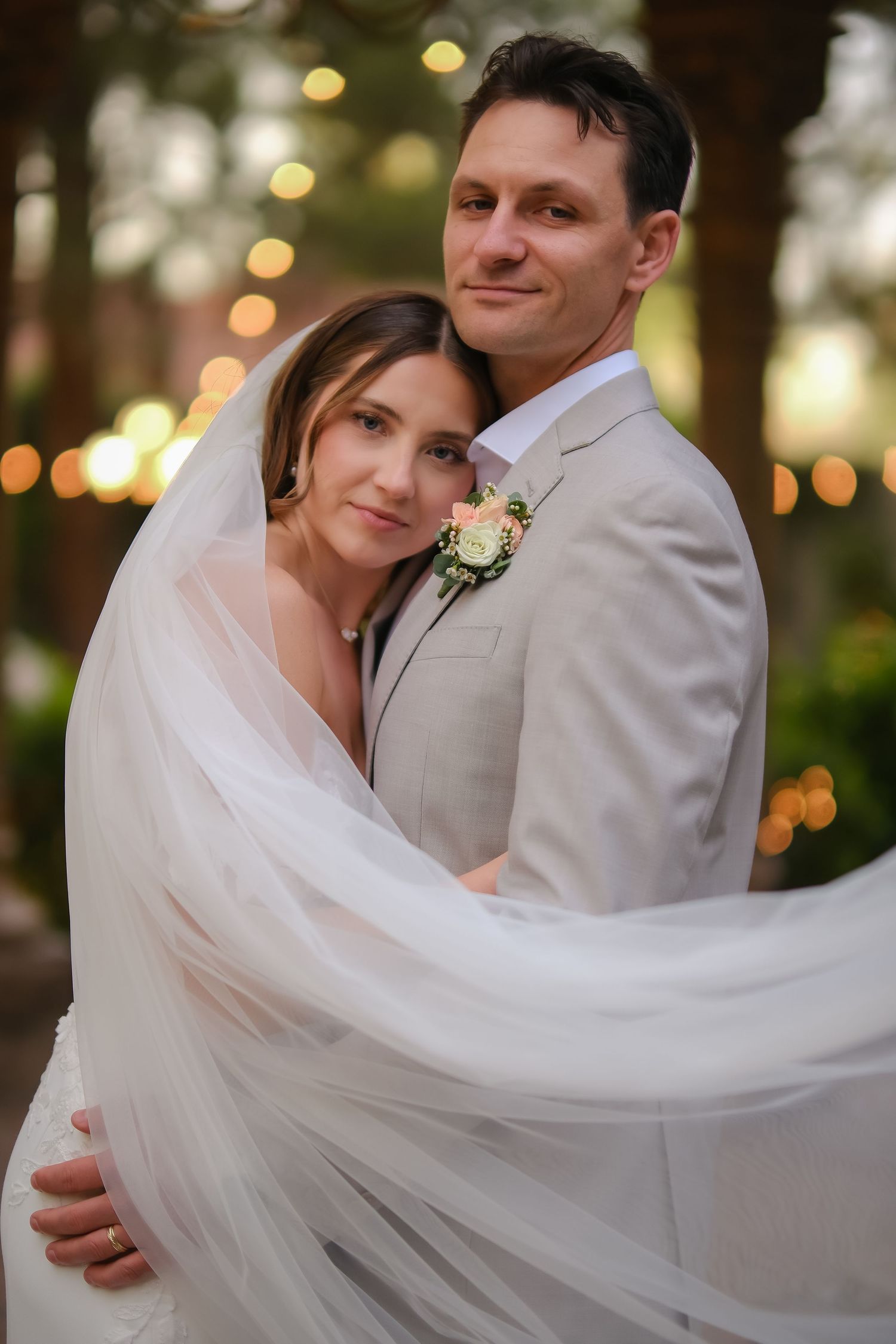 Bride in flowing veil embraces groom in tan suit with boutonniere, posing outdoors with warm bokeh lights behind them.