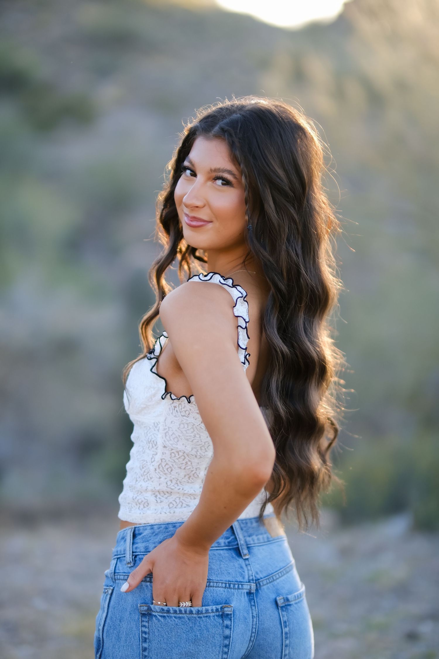 A person wearing a white lace top and denim jeans poses outdoors in natural lighting.