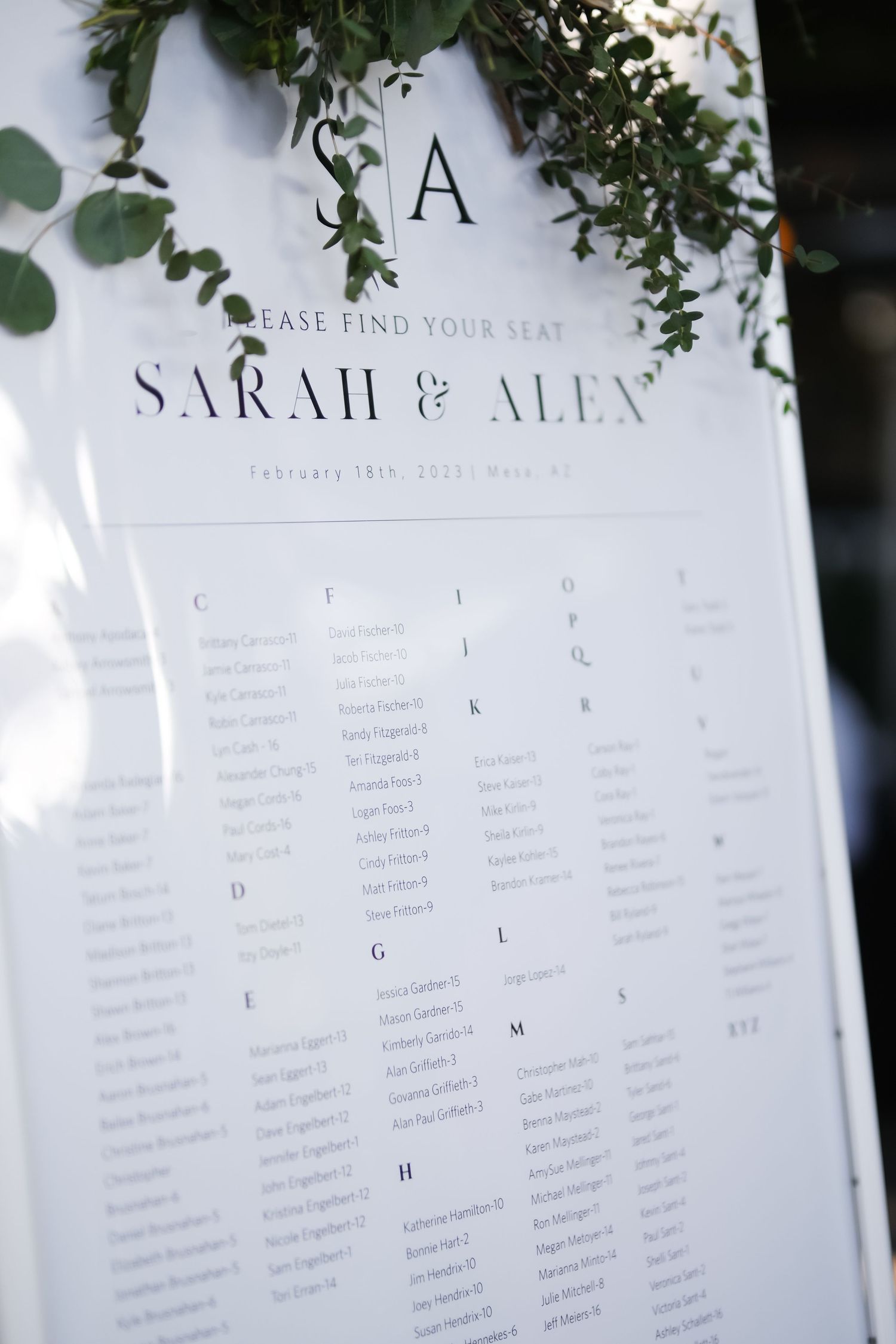 An elegant wedding seating chart with floral greenery decorations displays alphabetically organized guest table assignments.