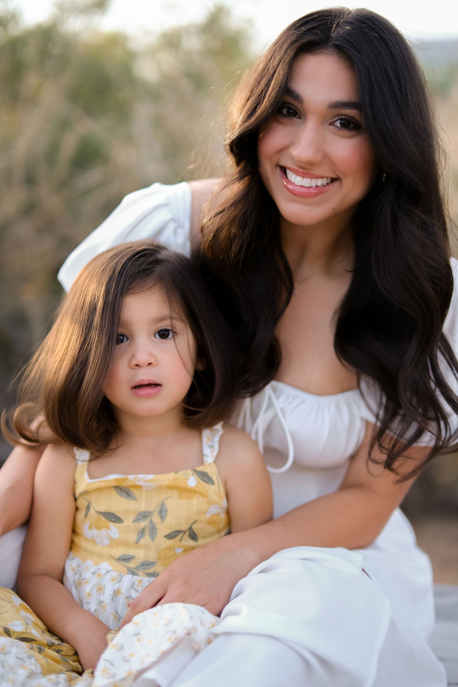 Mother and daughter in white and yellow sundresses pose together outdoors in warm natural lighting.