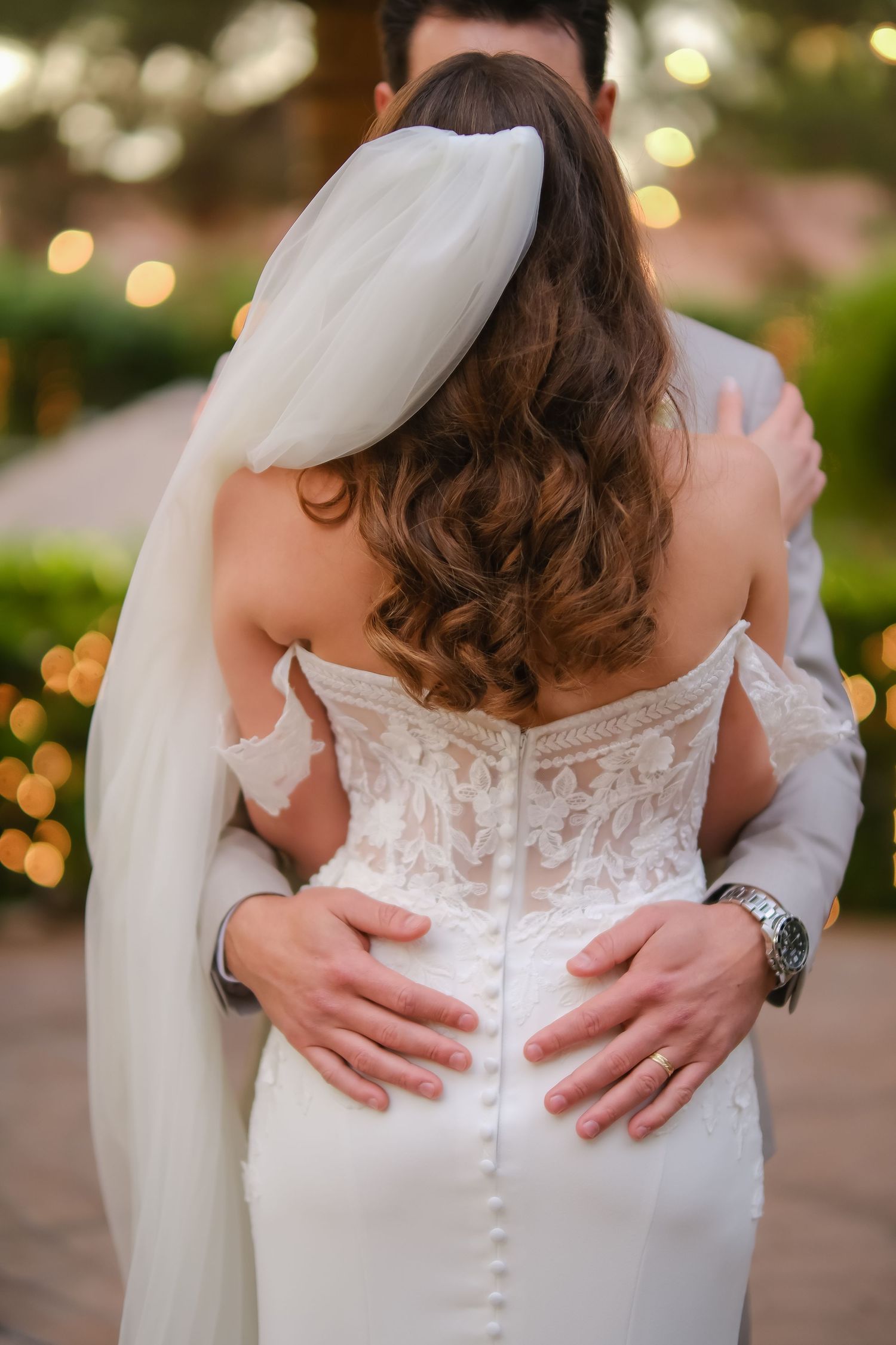 Bride and groom embracing, showing back of lace off-shoulder wedding dress with veil and button details.