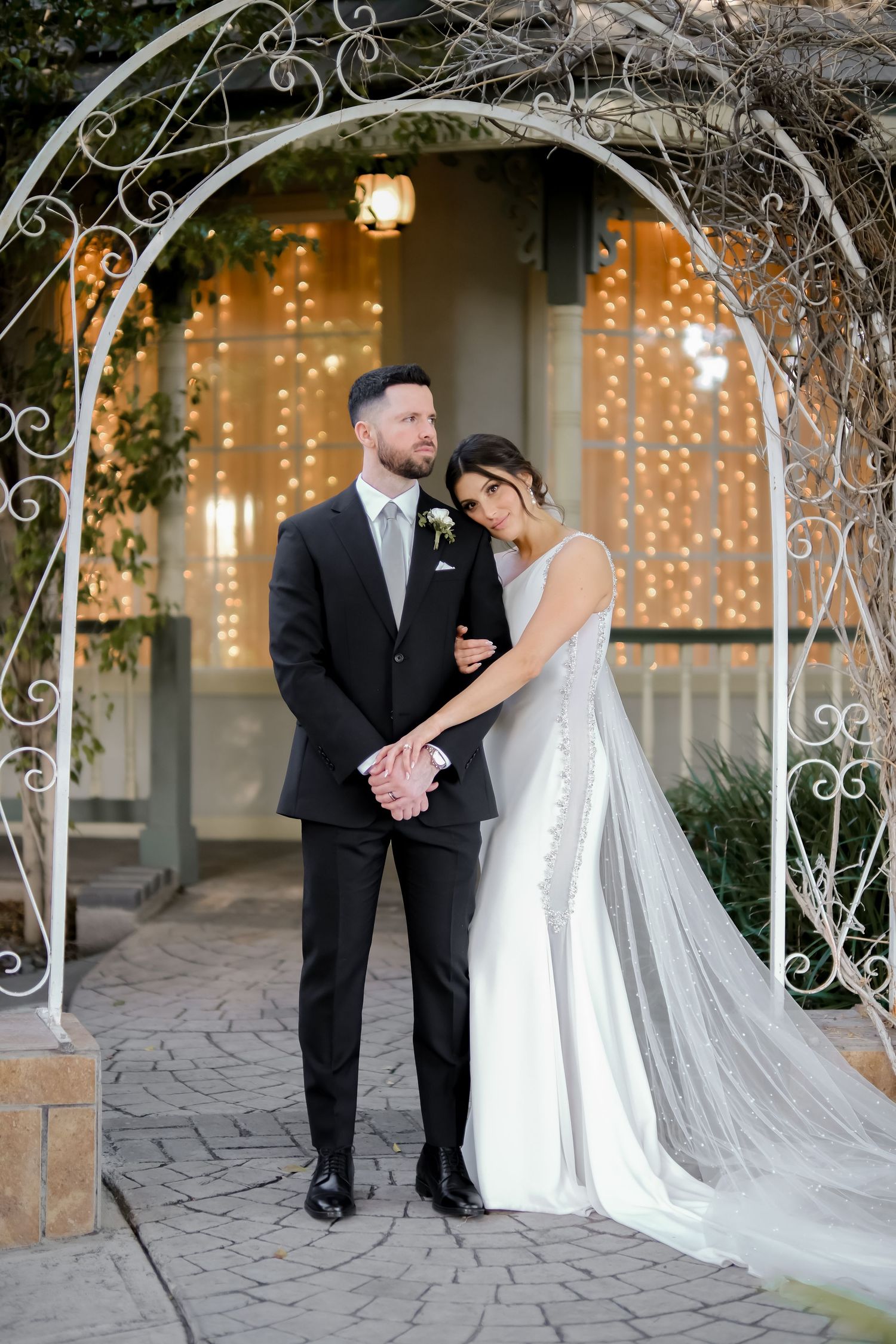 A wedding couple poses under an ornate metal arch with twinkling fairy lights in the background.