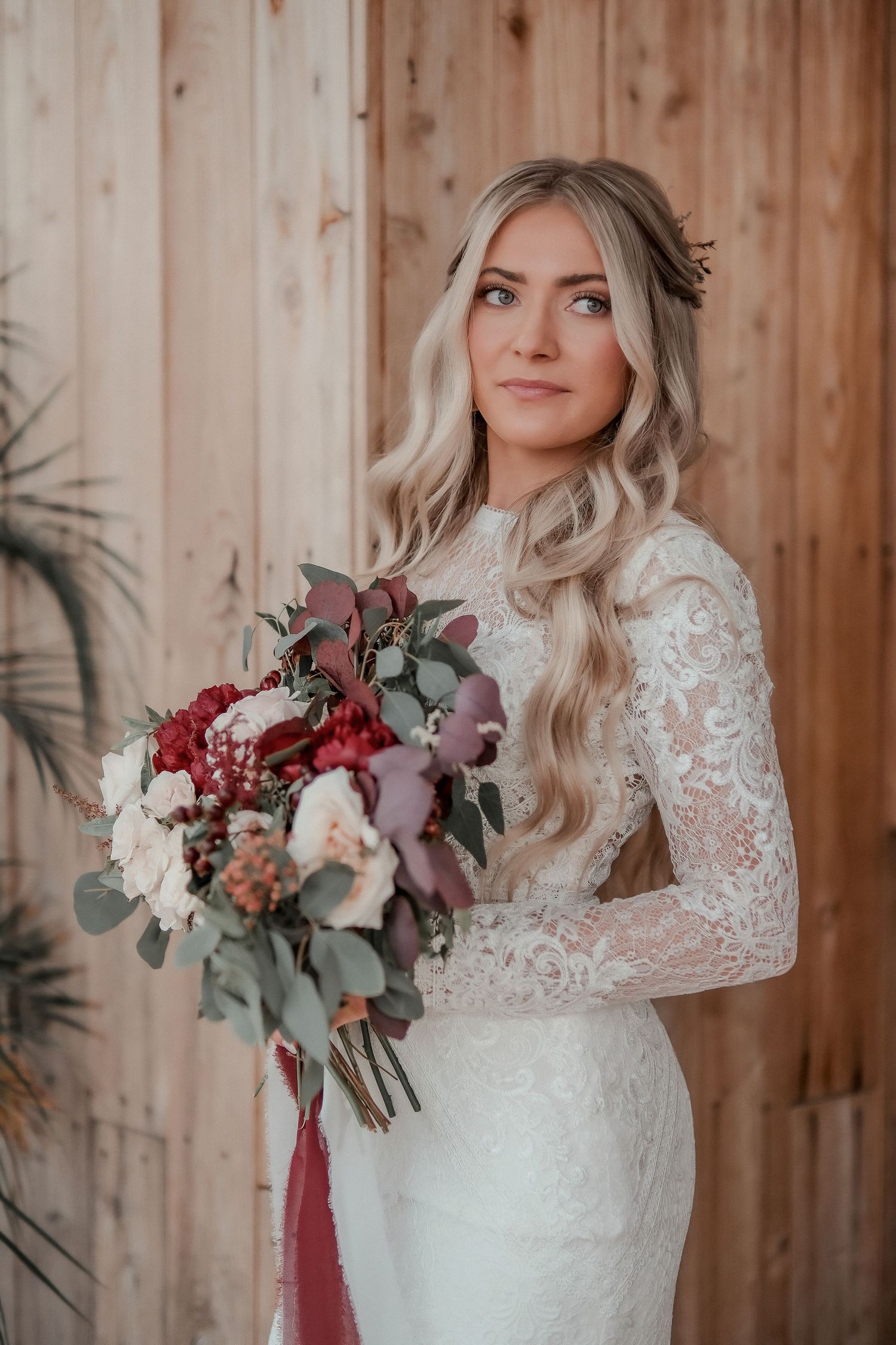 Bride in elegant lace wedding dress with long sleeves poses against rustic wooden wall with burgundy floral bouquet.
