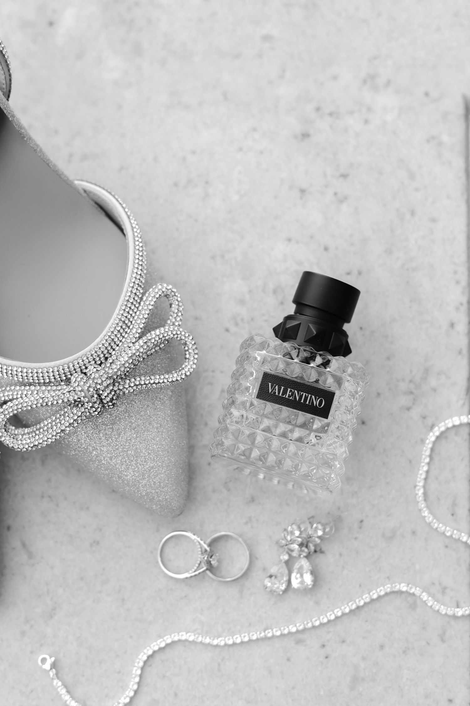 Flat lay of wedding accessories including rings, Valentino perfume, and crystal jewelry on light surface.