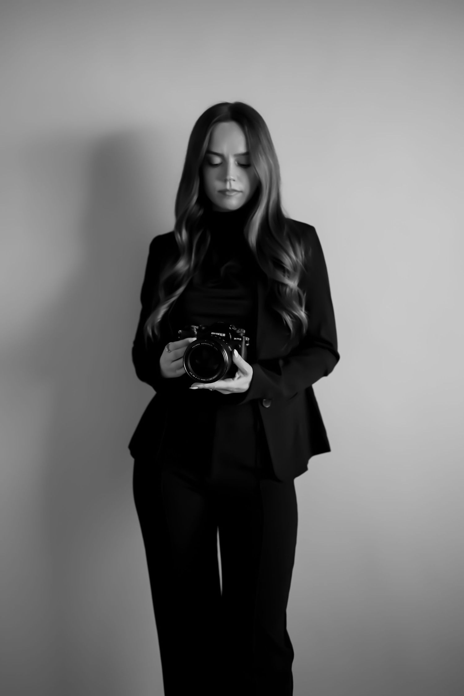 Black and white portrait of a photographer in an all-black outfit holding a camera while looking down.