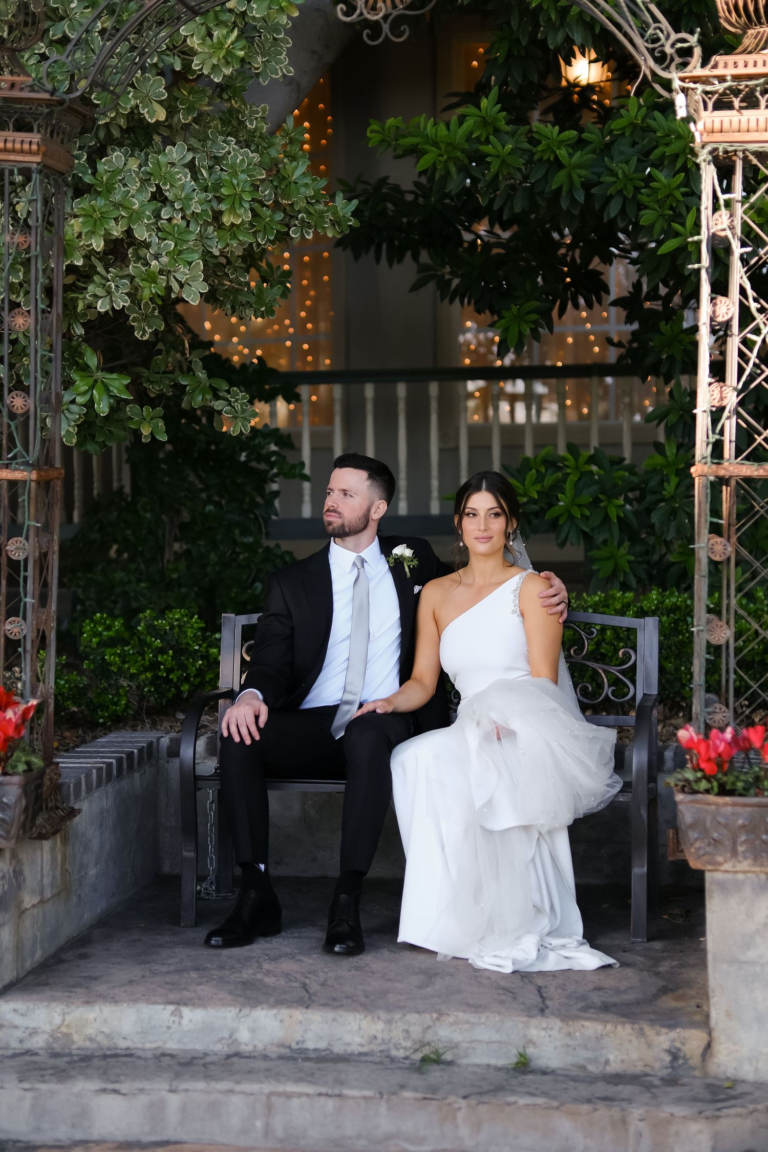 A couple in wedding attire sits on a bench under a decorative archway with string lights and greenery.