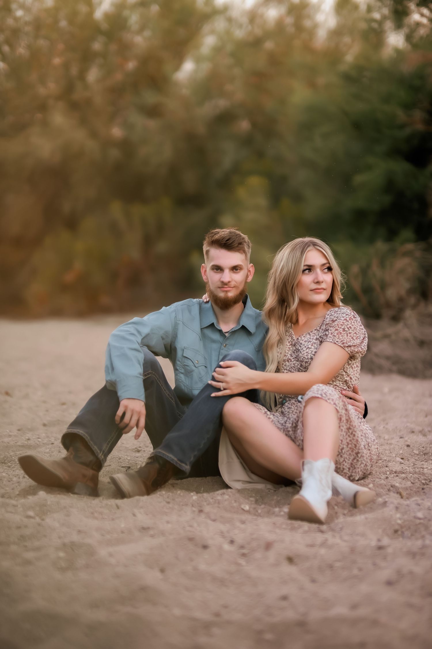 A casual couple sits together on sandy ground during golden hour photography session.