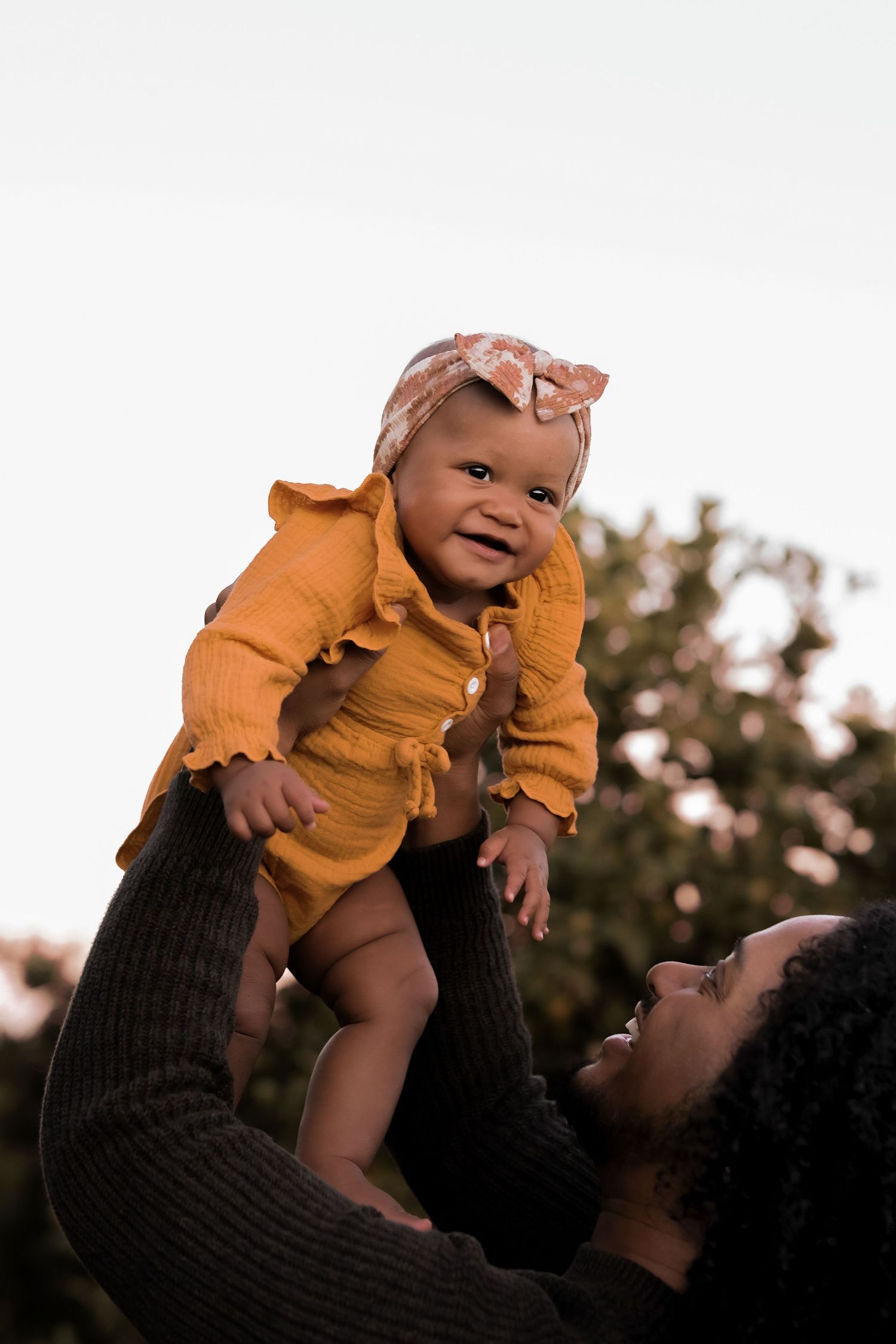 Baby in mustard hoodie being lifted by caregiver outdoors at sunset.